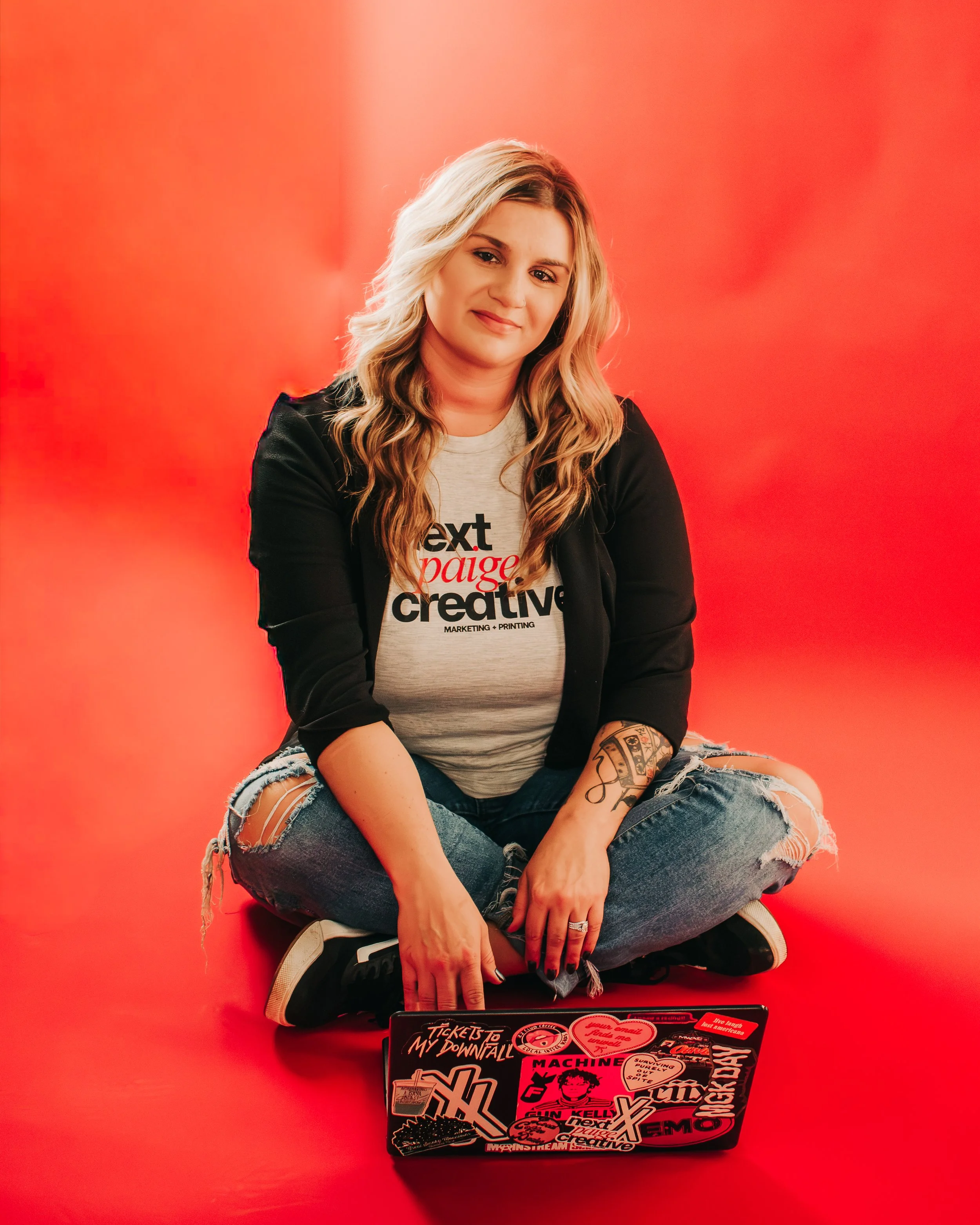 A young woman with long, wavy blonde hair wearing a light gray t-shirt with black, red, and white text, a black jacket, and ripped jeans, sitting cross-legged on a red background with her laptop covered in stickers in front of her.