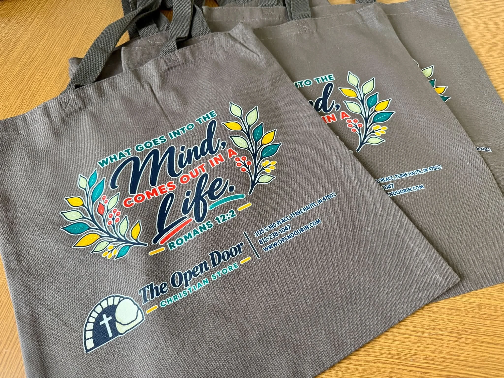 Multiple gray tote bags with a colorful inspirational quote and decorative leaves printed on them, laying on a wooden surface.