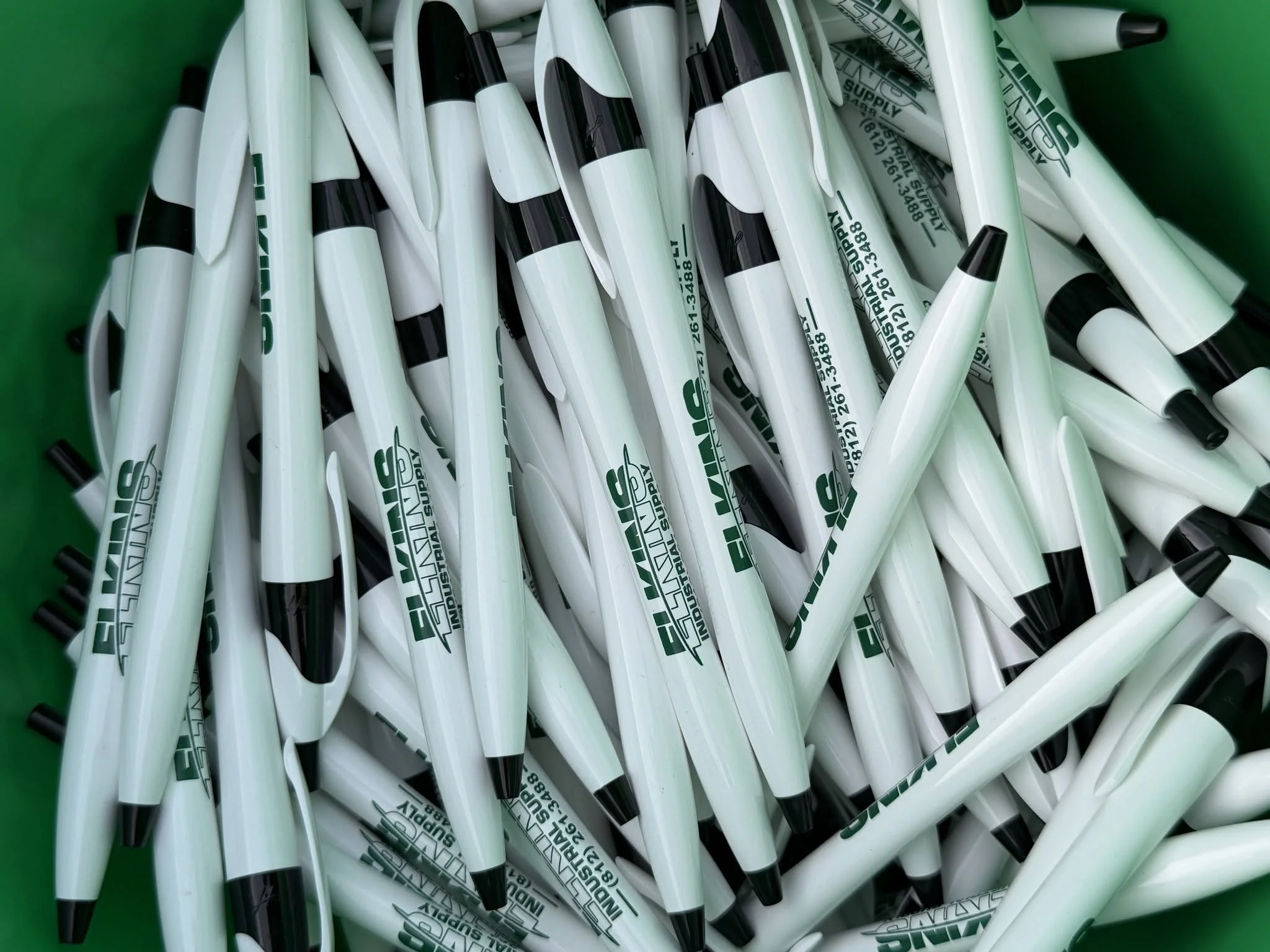 A pile of white and black marking pens with green text, inside a green container.