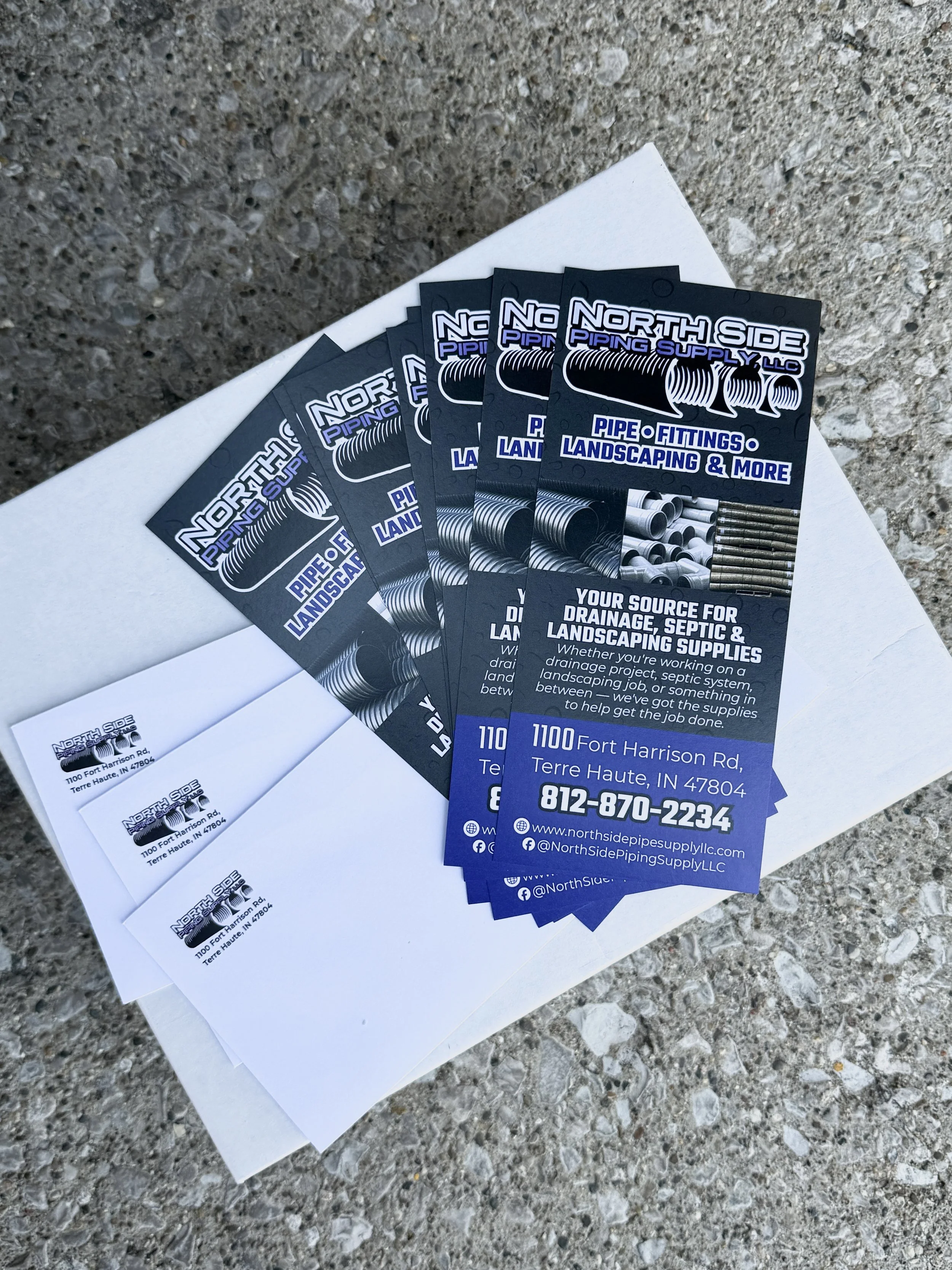 Multiple business flyers for North Side Piping Supply LLC are spread on a concrete surface, featuring information about pipe fittings, landscaping, plumbing, drainage, septic, and other supplies, with contact details included.