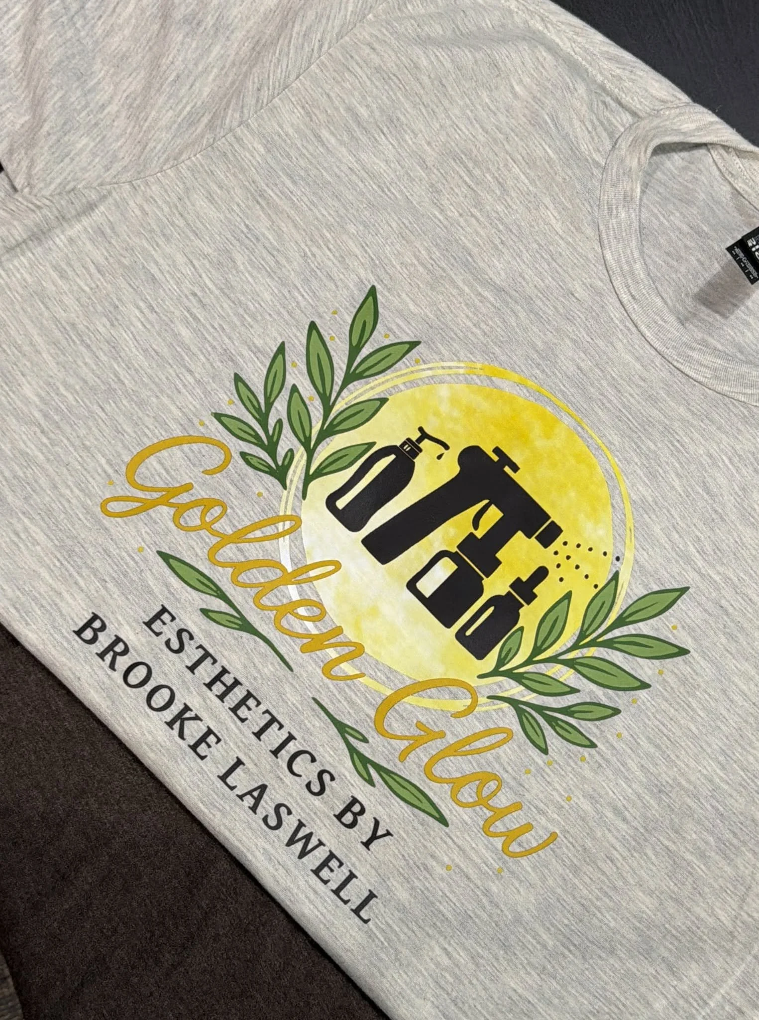 Close-up of a gray T-shirt with a gardening-themed design featuring a watering can, spray bottle, and green foliage, with the word 'Grow' in yellow script and other text related to organic and homemade baked goods and bakery.