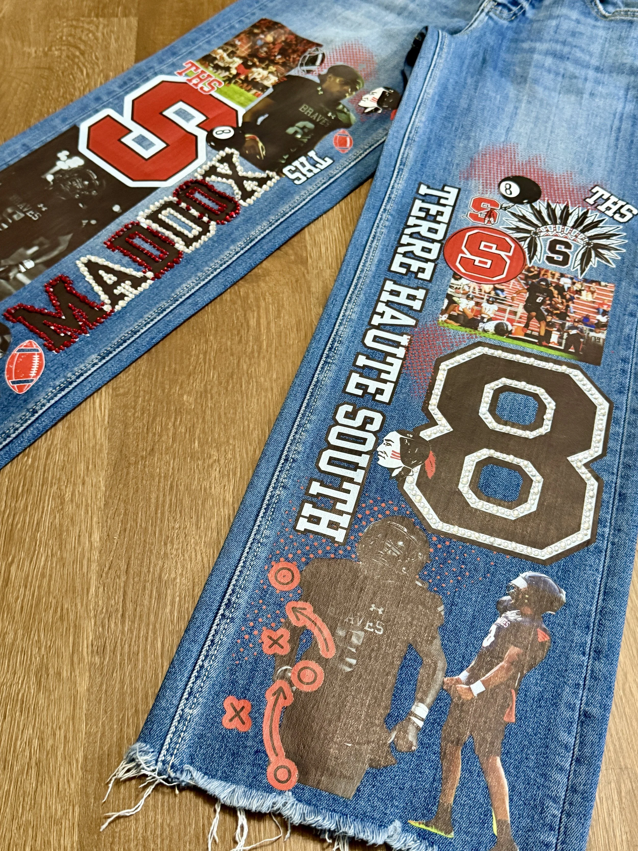 A pair of denim jeans decorated with sports-themed patches, stickers, and images, including a football player, the Ohio State University logo, and athletic motifs, laid out on a wooden surface.