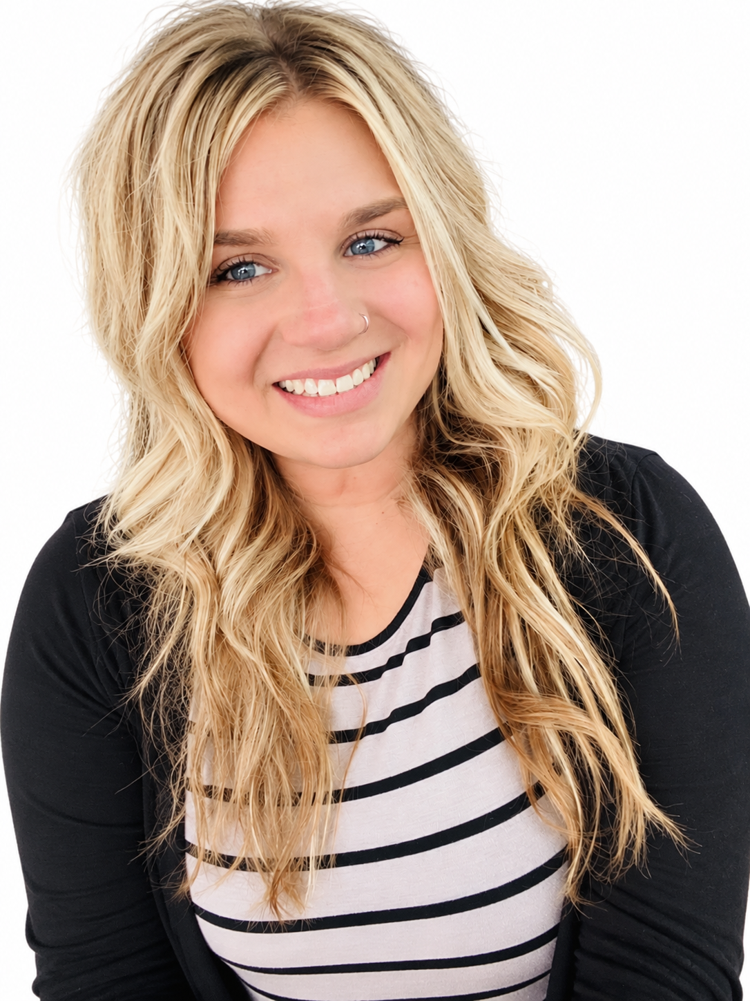 A young woman with long blonde wavy hair, blue eyes, and a nose piercing, smiling at the camera, wearing a black blazer over a white and black striped shirt against a white background.