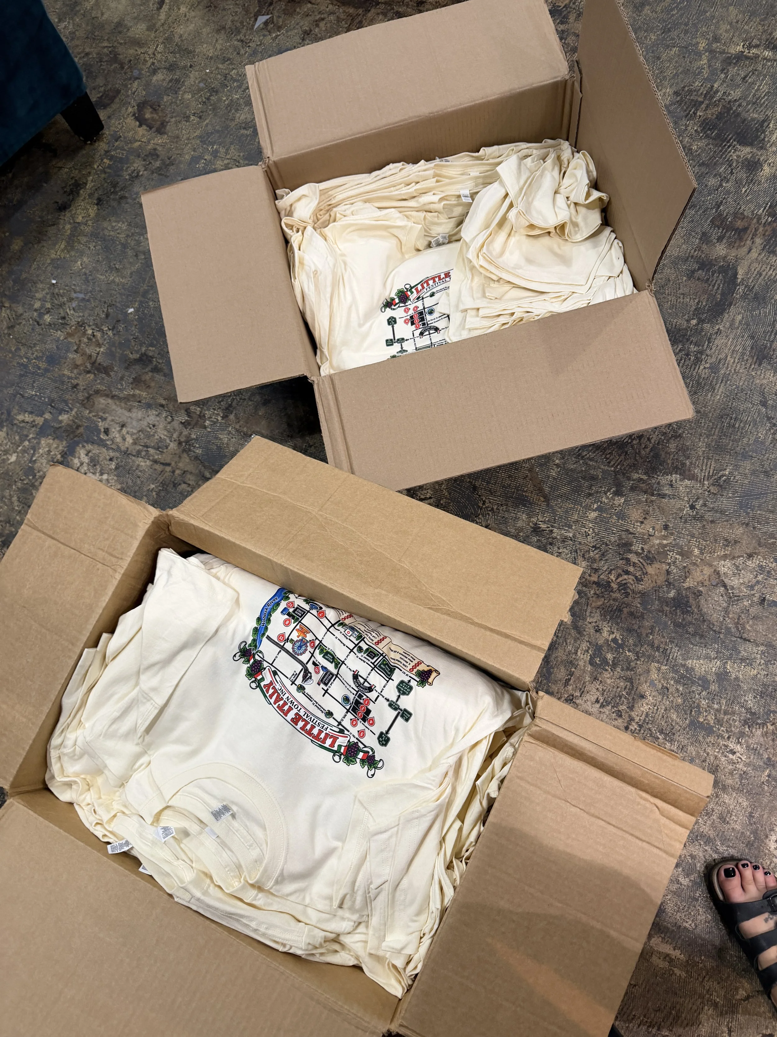 A cardboard box filled with beige T-shirts featuring a colorful graphic design, placed on a rough wooden floor. A person's foot with a sandal is visible in the corner.