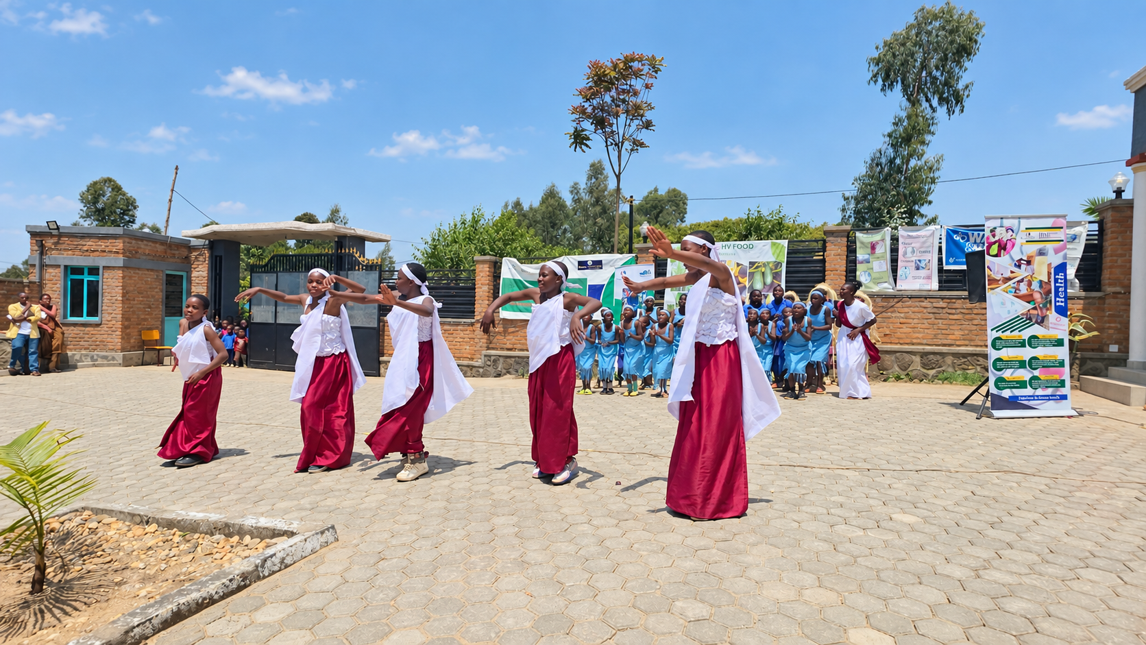 Girls perform a dance in front of an audience during an outdoor event on a bright, sunny day with a blue sky. They are dressed in red skirts and white tops, and some in white headbands. Behind them, other children and adults watch, with banners and posters displayed along the brick wall.