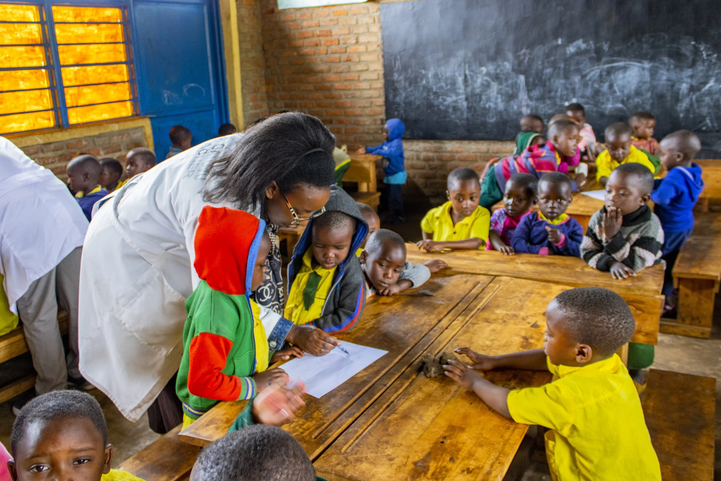 An adult works with a child at a classroom desk while other children sit nearby.