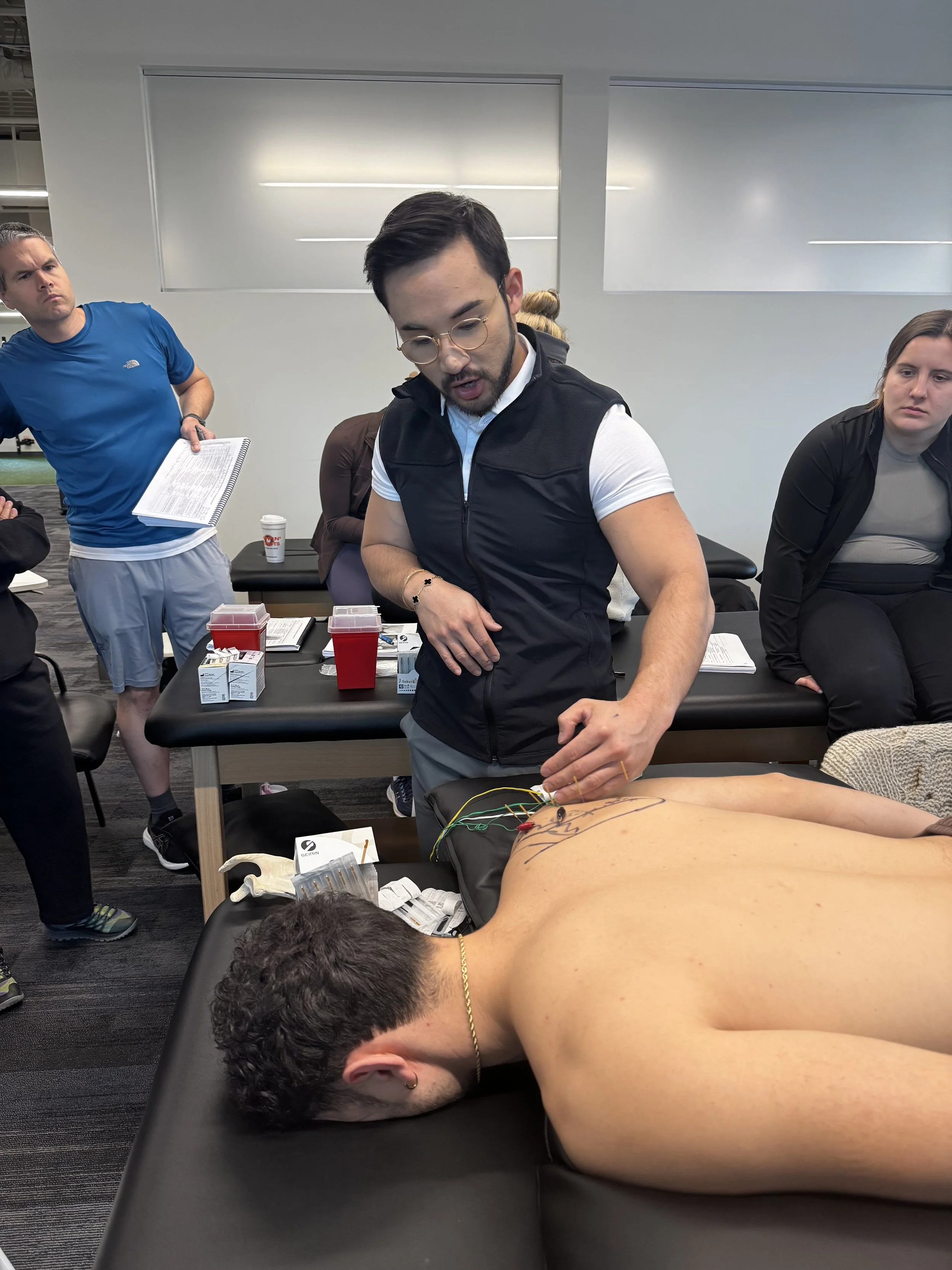 A person lying face down on a medical table with wires attached to their back, during a medical procedure or training session, with several people observing or assisting.