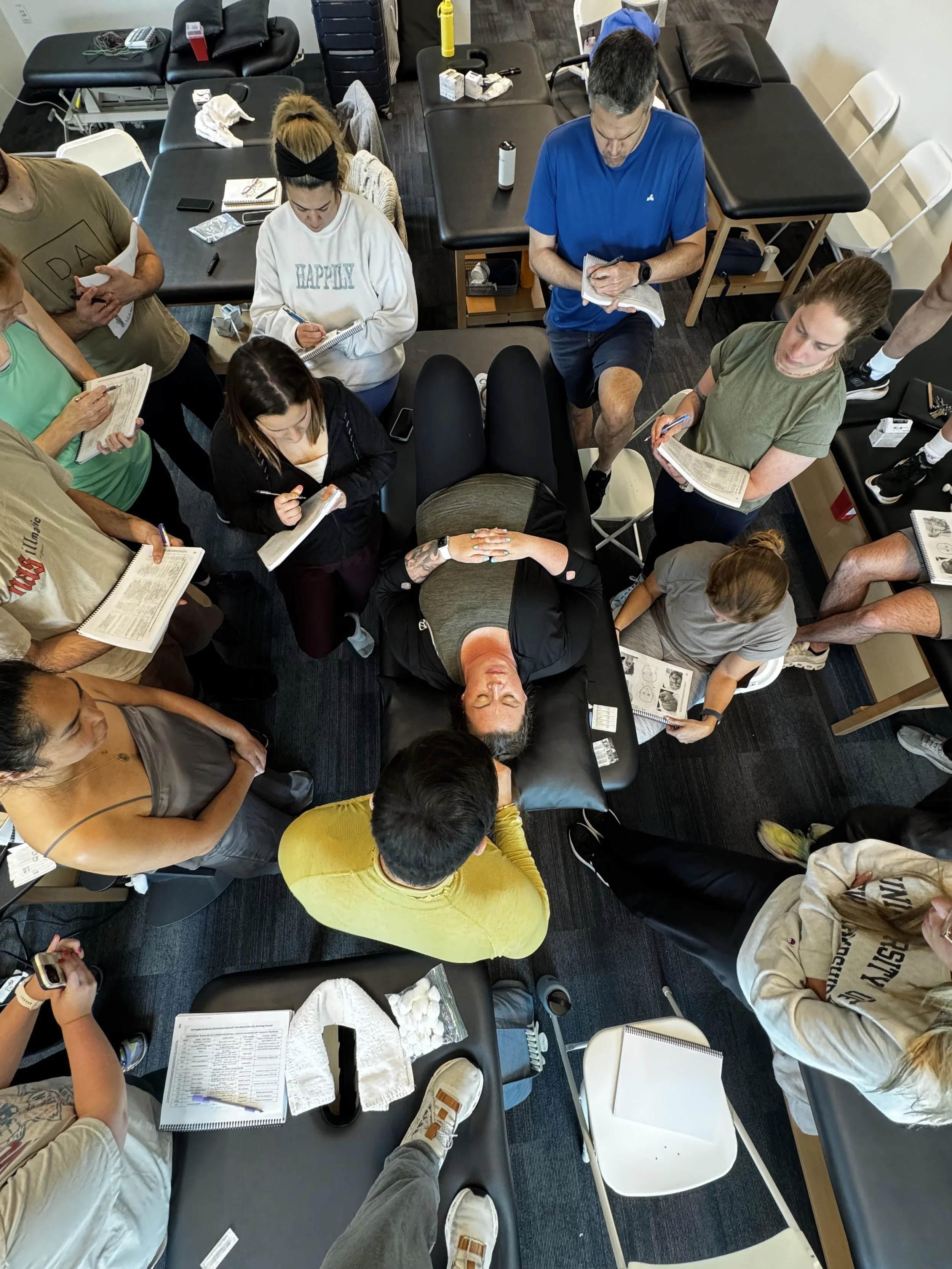 Overhead view of a group of people gathered around tables in a fitness or therapy setting, some taking notes, others resting or talking, with fitness equipment and supplies on the tables.
