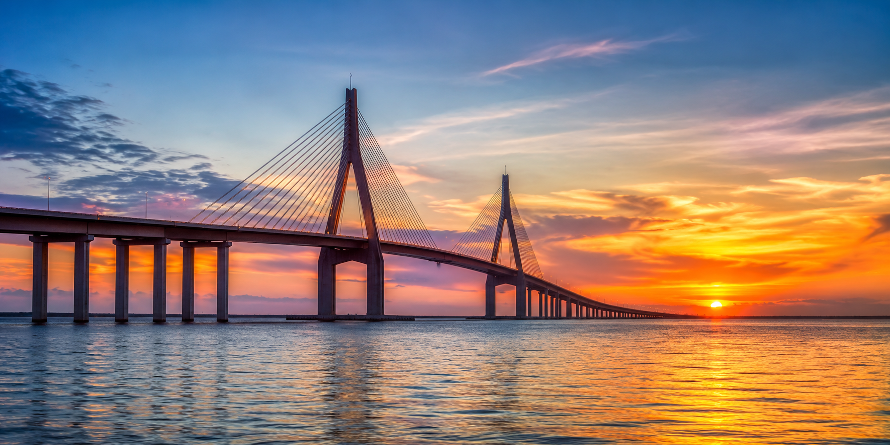 The Sunshine Skyway bridge extending over Tampa Bay at sunset with the sun near the horizon, colorful sky with clouds, and reflections on the water.