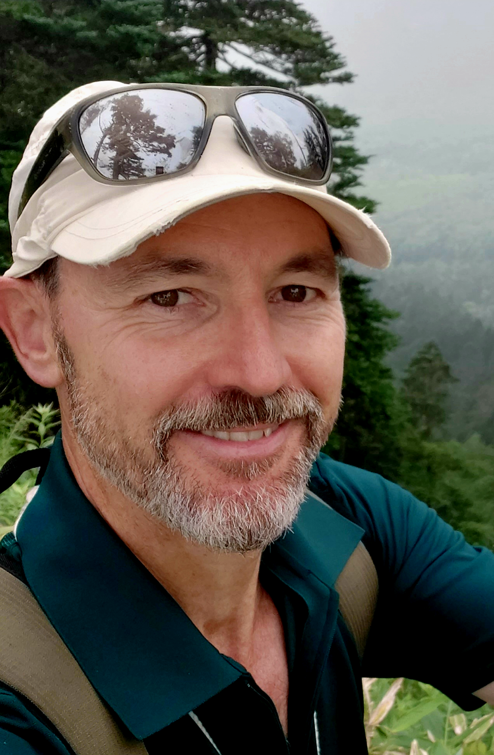 A smiling man with a beard and sunglasses on his white cap, outdoors with green trees and foggy weather in the background.