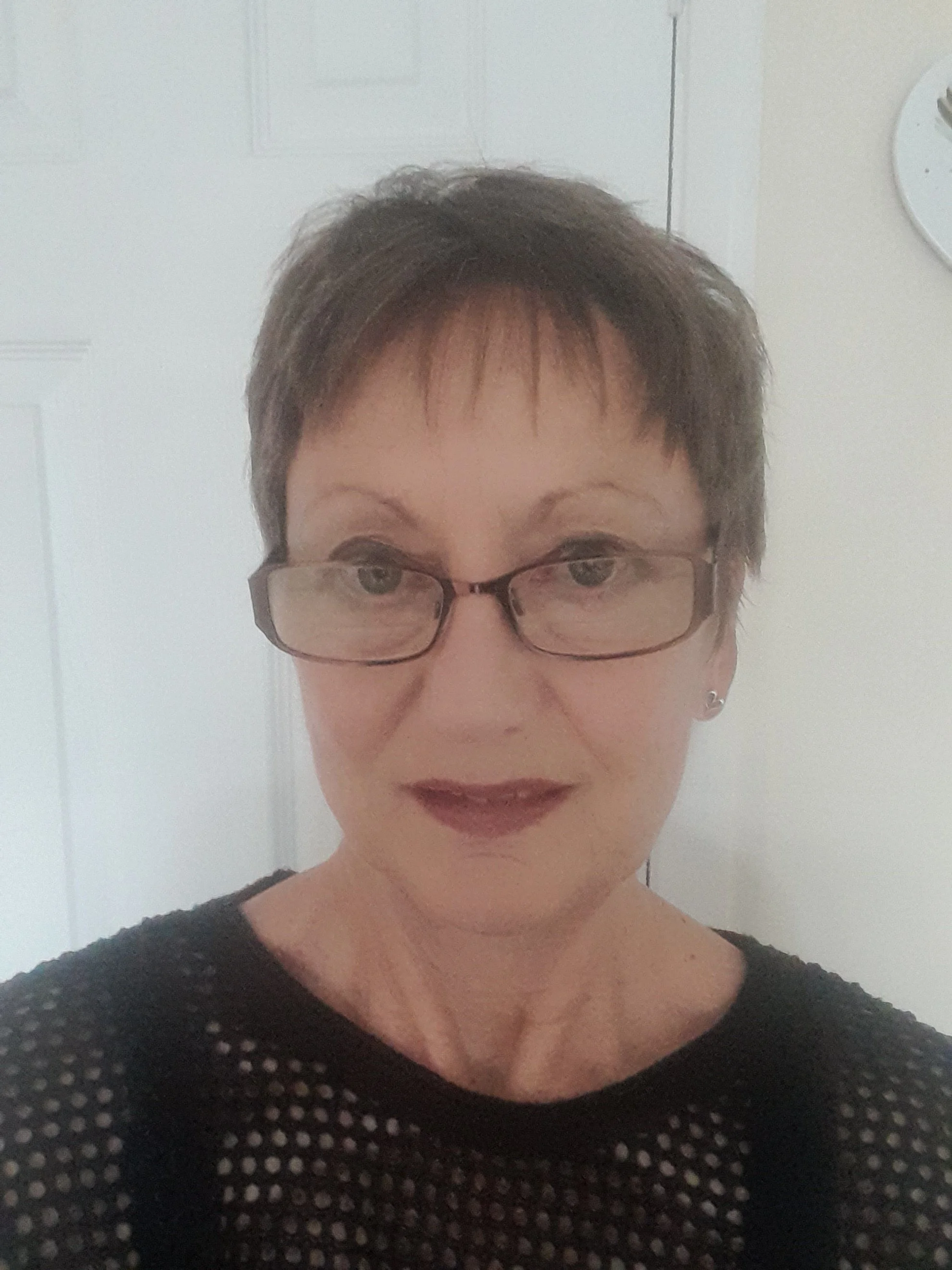 A woman with short dark hair and glasses, wearing a black mesh top, standing indoors against a white door.