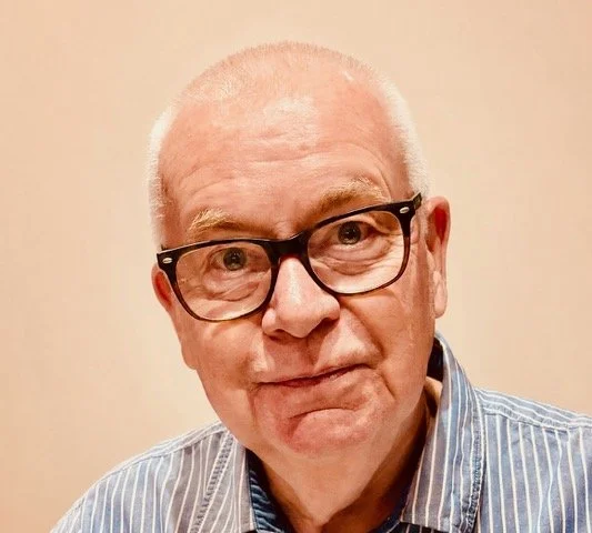 A close-up of an elderly man wearing glasses and a striped shirt, with a neutral background.