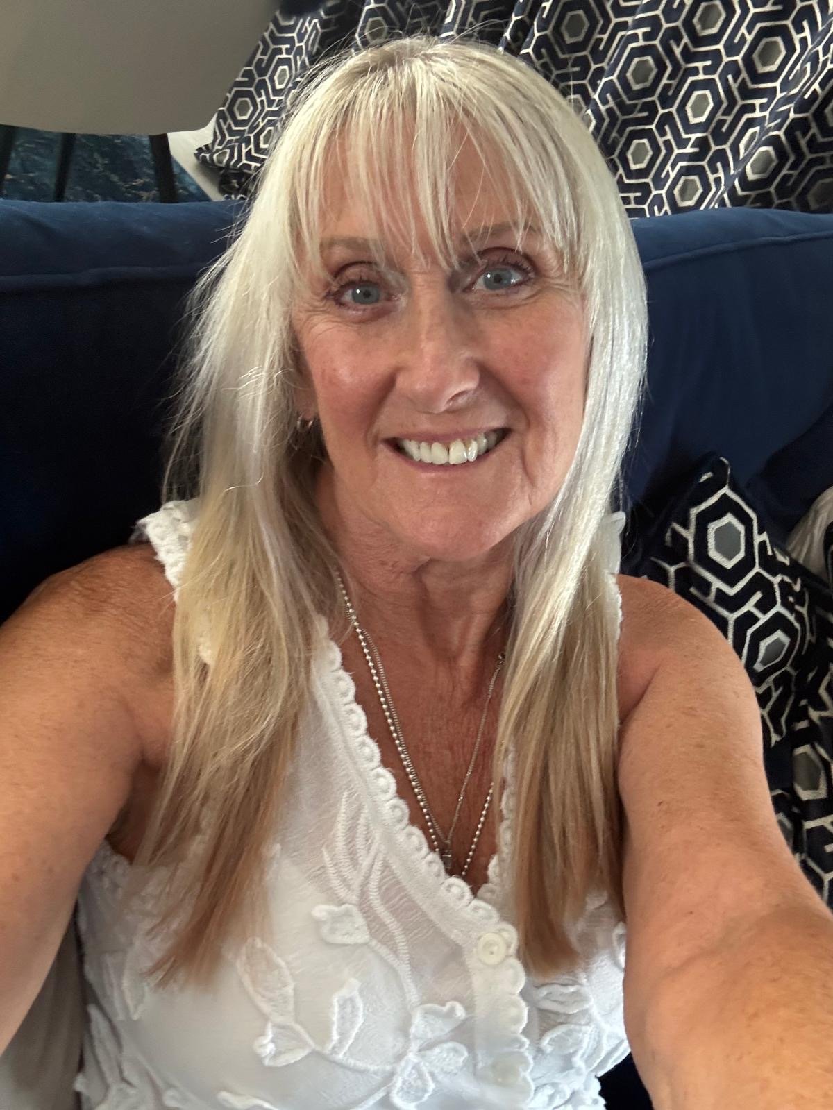 A smiling middle-aged woman with long blonde hair sitting on a sofa, wearing a white sleeveless top with lace embroidery, necklaces, and earrings. There are patterned curtains and a dark cushion in the background.
