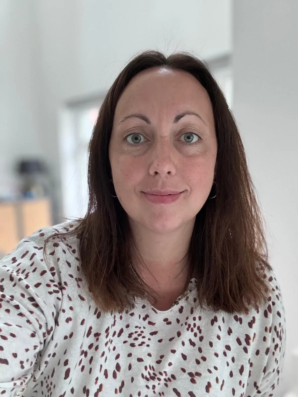 Close-up of a woman with shoulder-length brown hair and light blue eyes, wearing a white shirt with red spots, smiling in a bright indoor setting.