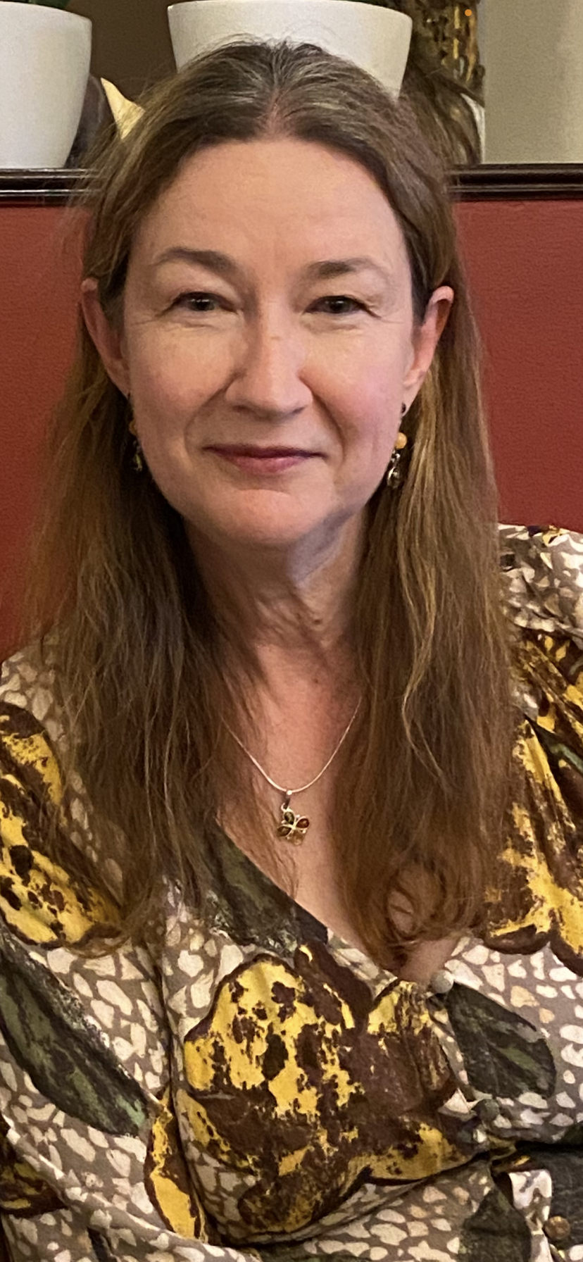 A woman with long brown hair and jewelry, wearing a patterned dress, seated indoors against a reddish background.