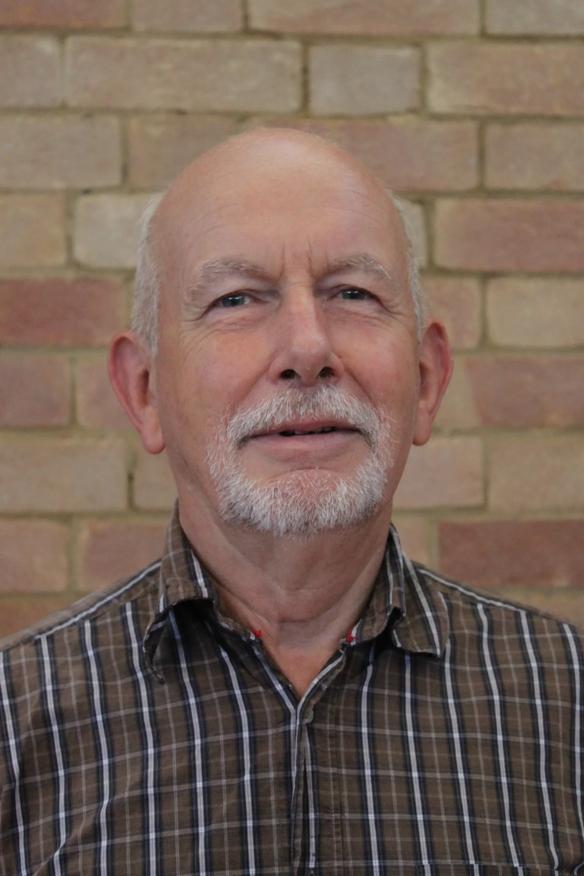 Close-up of an older man with a mustache and beard, wearing a plaid shirt, standing in front of a brick wall.