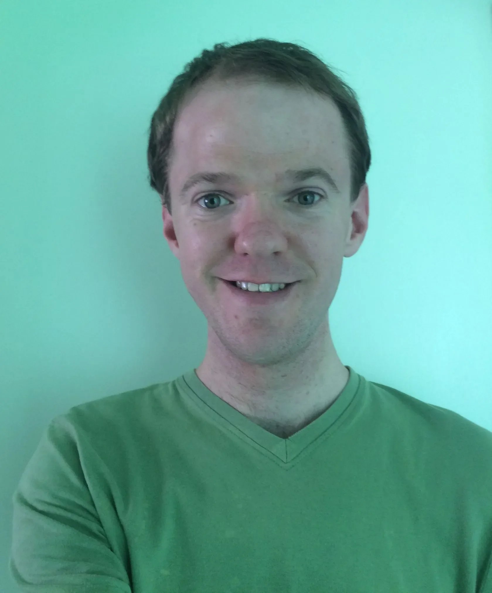 Close-up of a man with blue eyes, short brown hair, and a light green shirt, smiling in front of a plain teal background.
