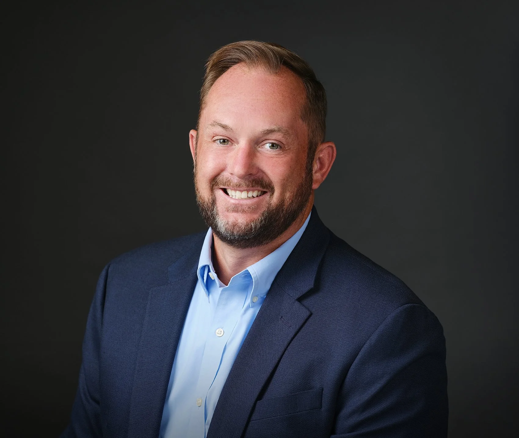 A professional headshot of a smiling man with a beard and short brown hair, wearing a navy blue blazer and light blue dress shirt, against a dark background.
