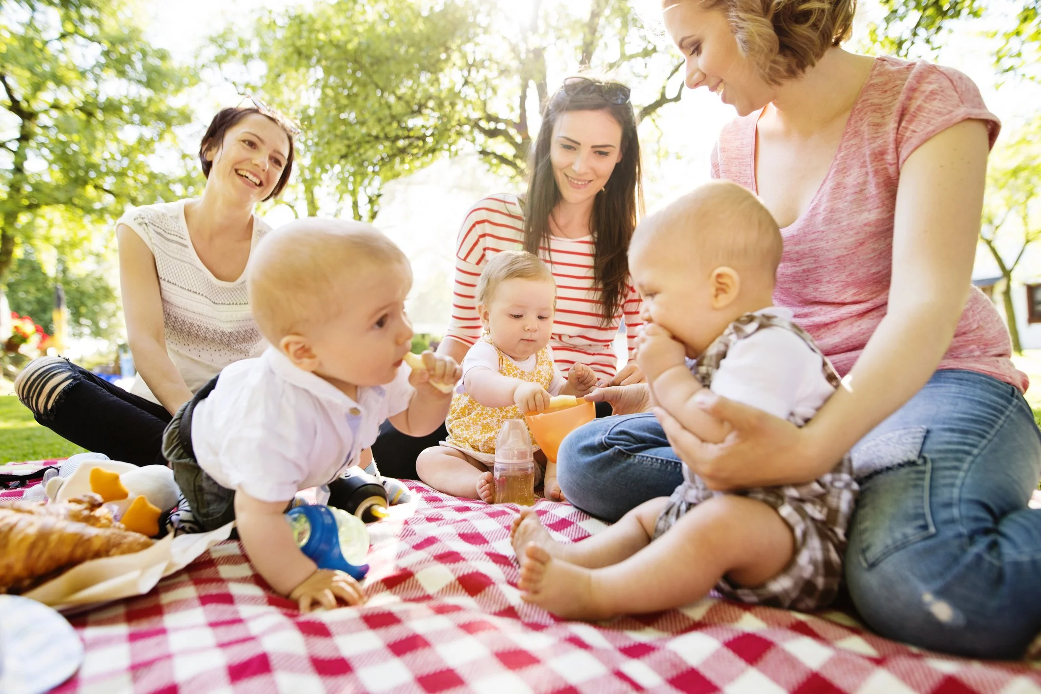 Group of women and young children having a picnic outdoors on a sunny day, sitting on a red and white checkered blanket with food and toys.