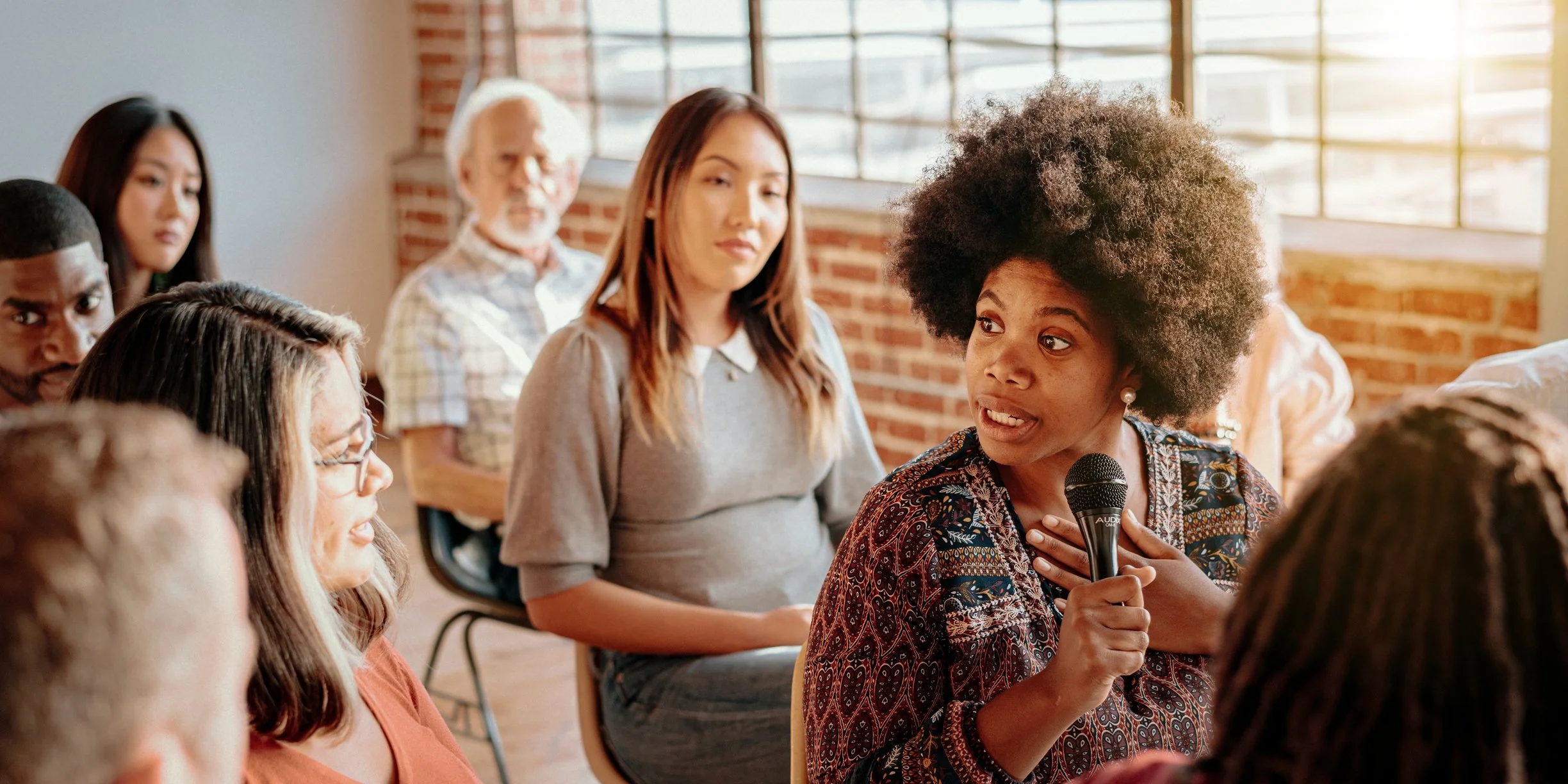 Woman with an Afro hairstyle holding a microphone speaking during a group discussion in a bright room with exposed brick wall and large windows.