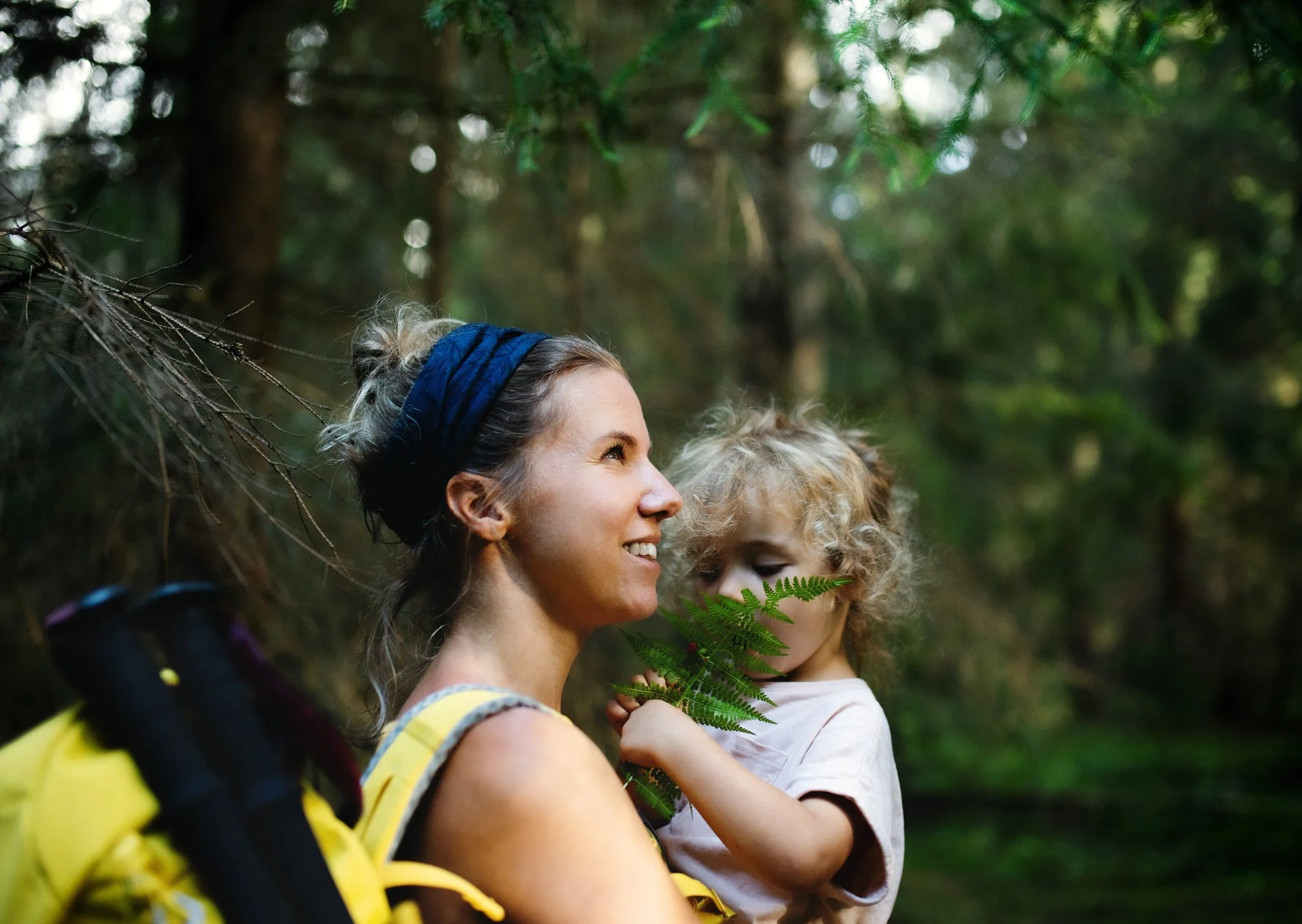 A woman with a black headband smiling and holding a young girl with curly hair, surrounded by trees and greenery in a forest.