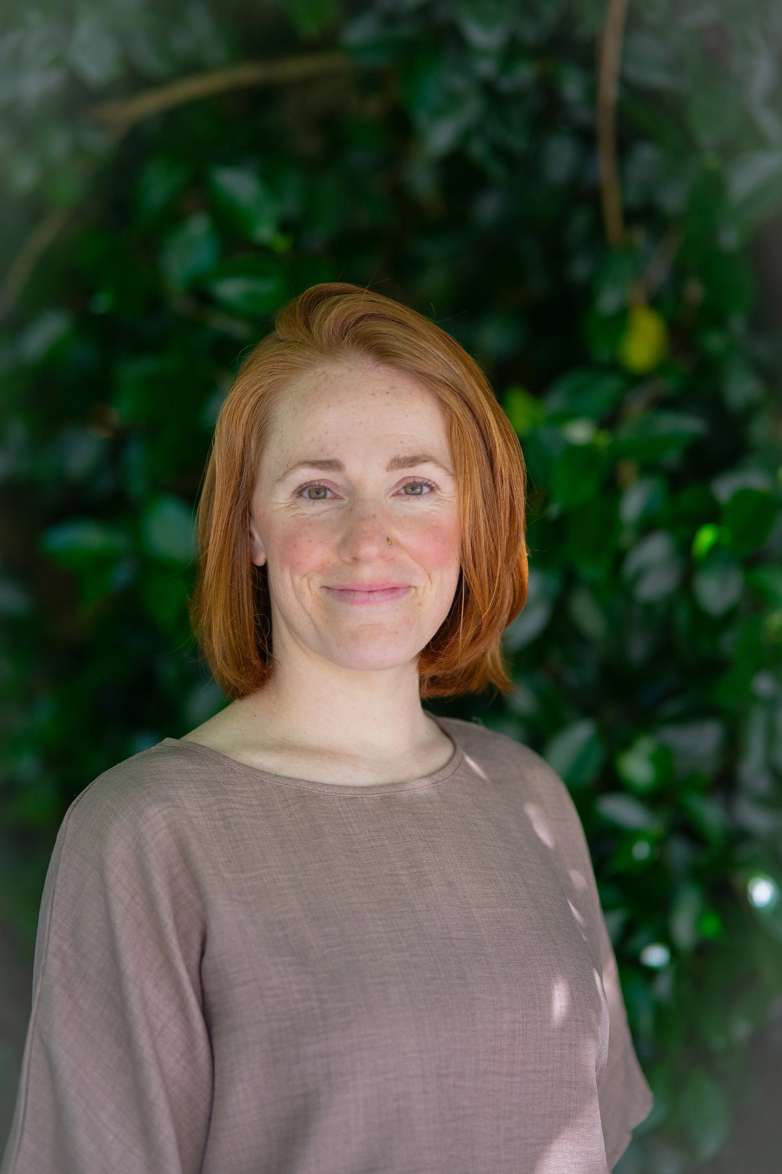 A woman with red hair and a nose piercing smiling, standing outdoors in front of green foliage wearing a light brown long-sleeve top.