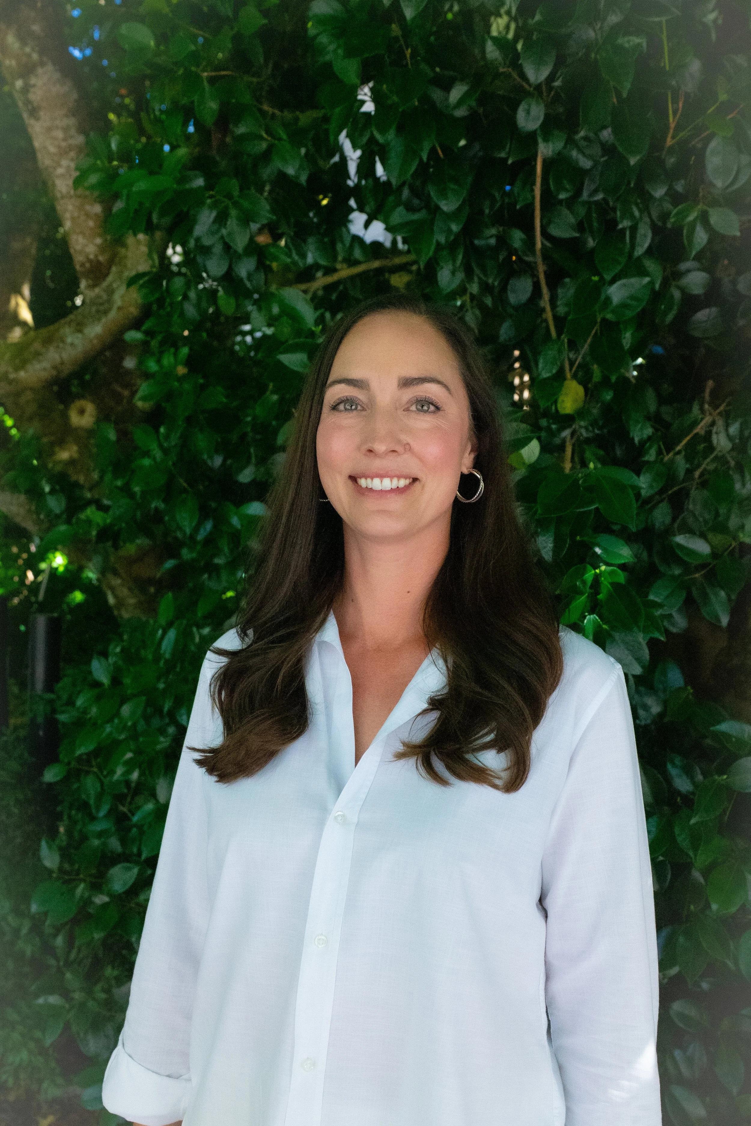 A woman with long brown hair and hoop earrings smiling and wearing a white shirt, standing in front of leafy green foliage.