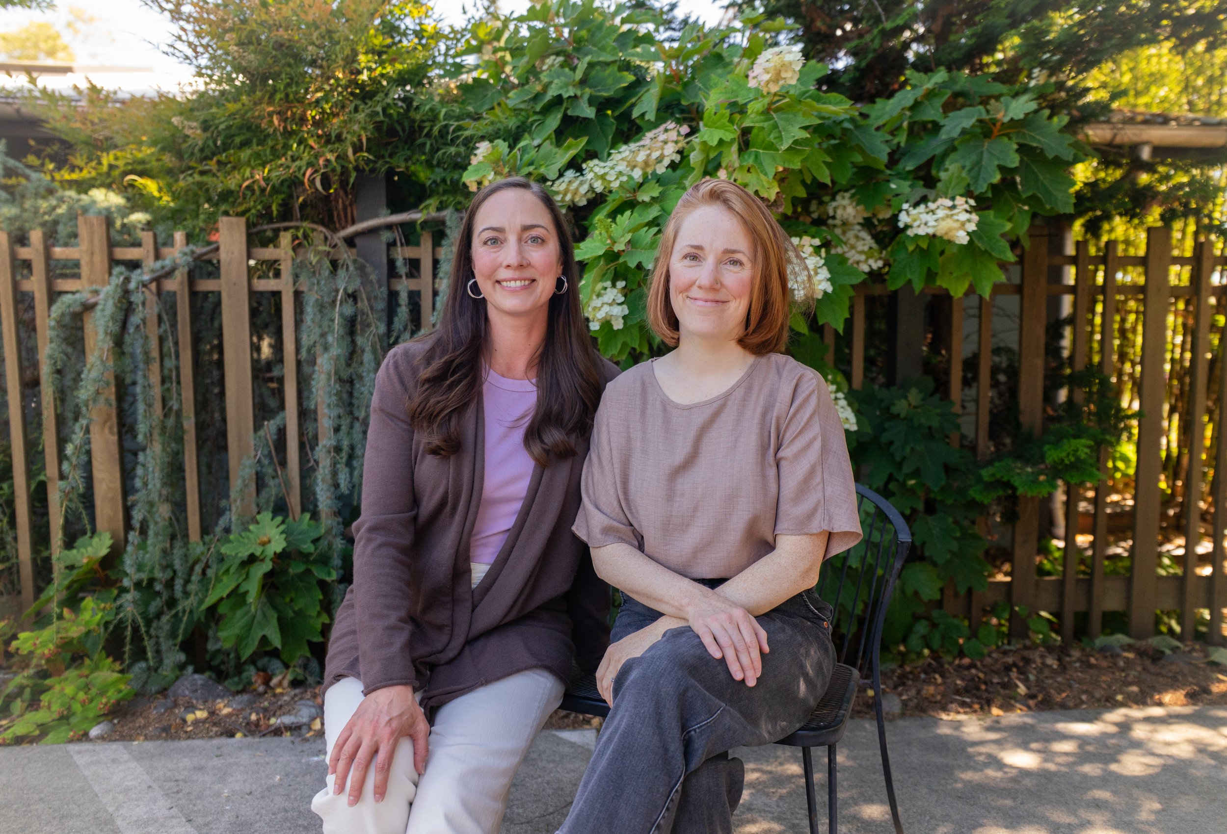 Two women under a large leafy tree with white flowers, sitting on a bench in a garden or park.