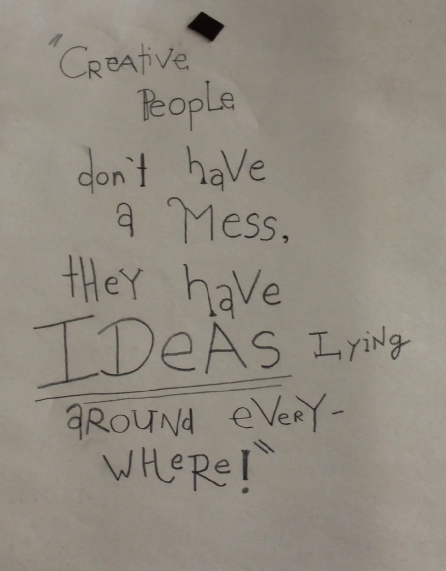 A handwritten quote on a white wall that reads, 'Creative People don't have a Mess, they have IDEAS lying around every WHERE!'