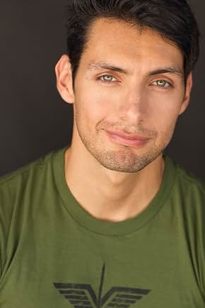 Close-up of a young man with dark hair and green eyes, wearing a green shirt, smiling subtly against a dark background.