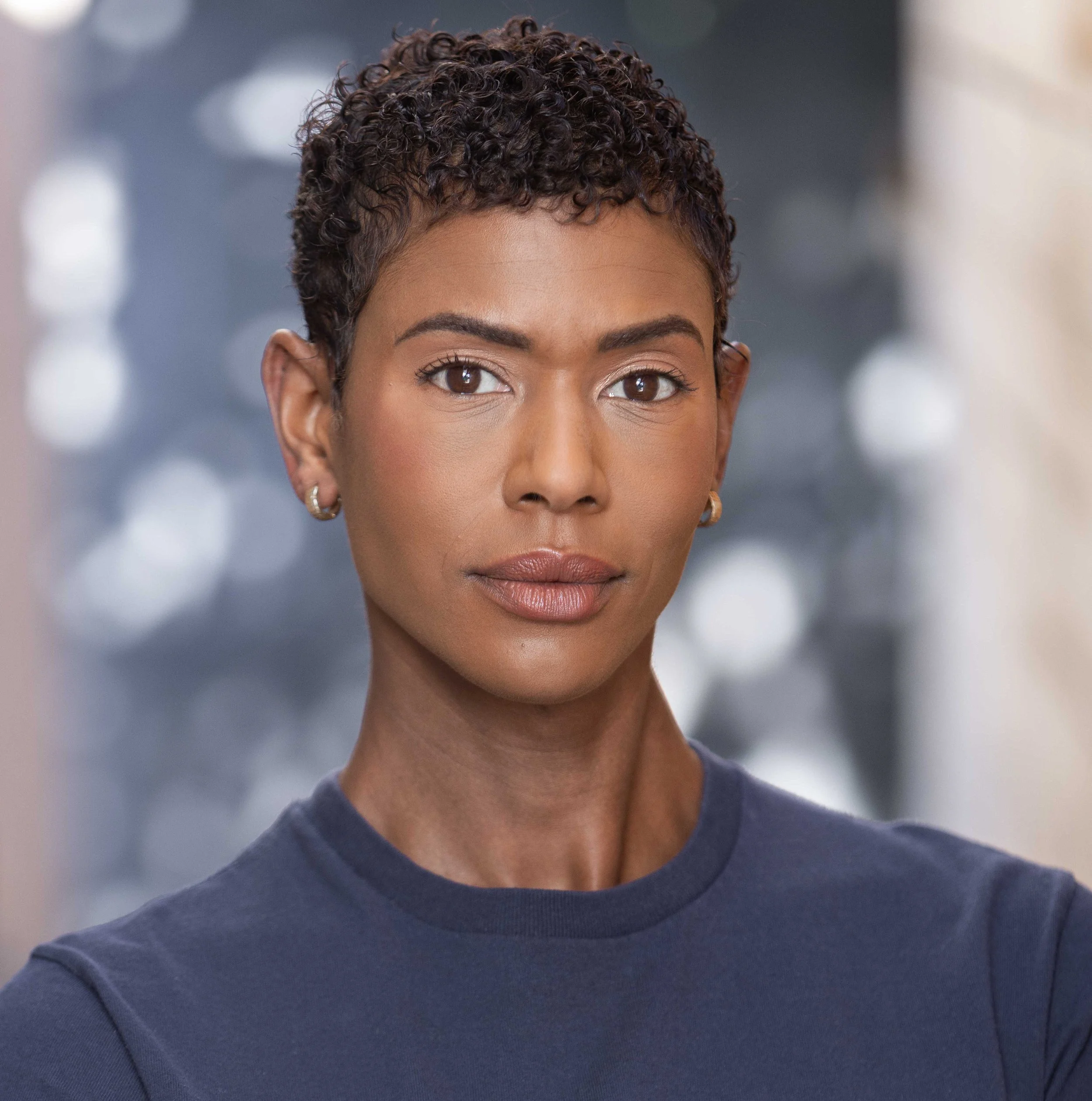 Close-up of a woman with short, curly hair and brown eyes, wearing a dark blue shirt and earrings.