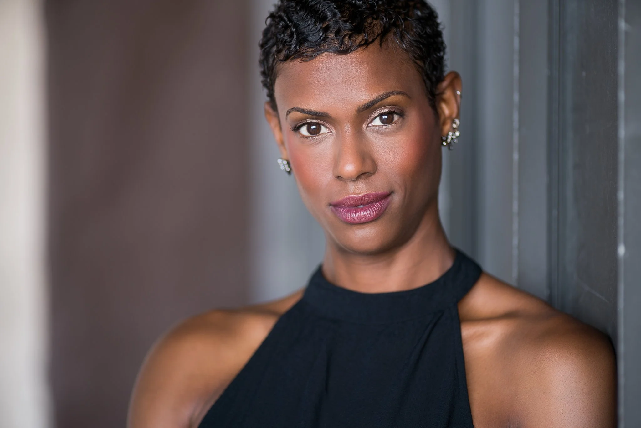 A close-up portrait of a woman with short, curly black hair, brown eyes, and light makeup, standing indoors near a dark-colored wall, wearing a sleeveless black top.