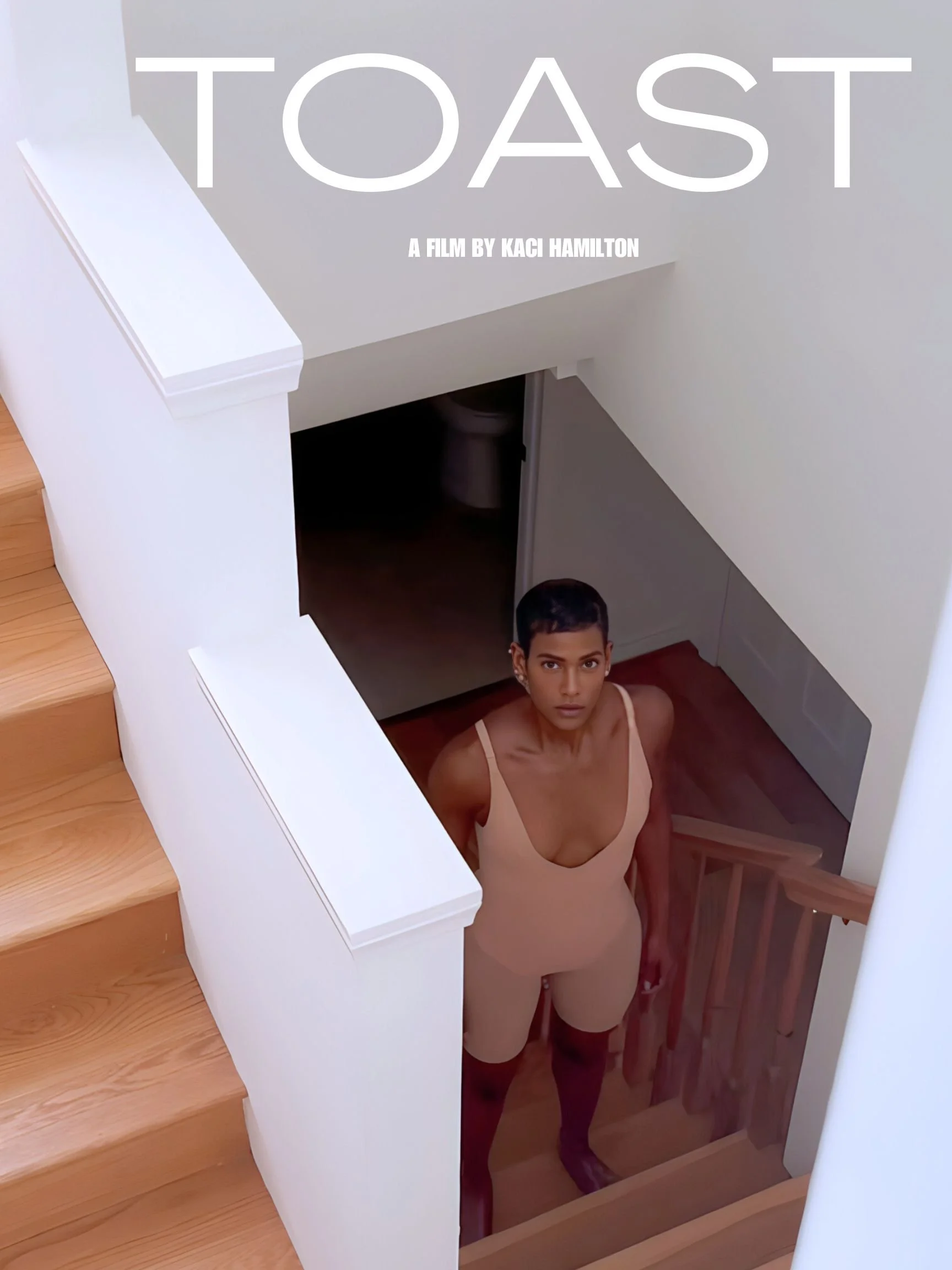 A woman standing at the bottom of stairs looking up. She has dark hair, is wearing a light-colored tank top, and black knee-high socks.