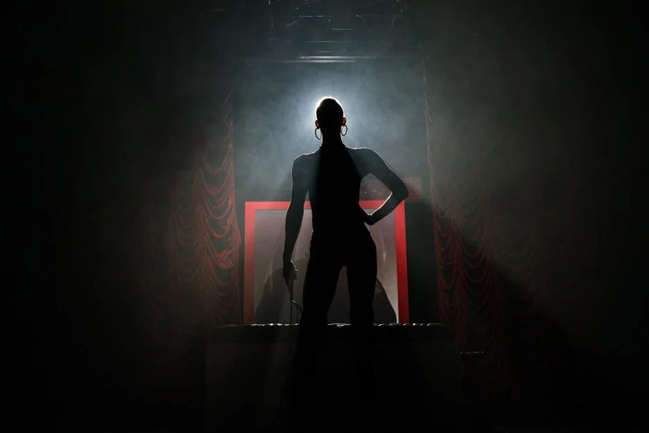 A silhouette of a woman standing on a stage, backlit with a bright light, holding a microphone in her right hand, against a backdrop with red curtains.