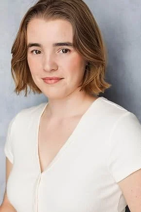 A young woman with short, light brown hair and fair skin, wearing a white top, smiling softly against a plain background.
