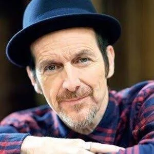 Close-up of a man with a beard and mustache wearing a dark blue hat and a plaid shirt, smiling slightly, outdoors setting.