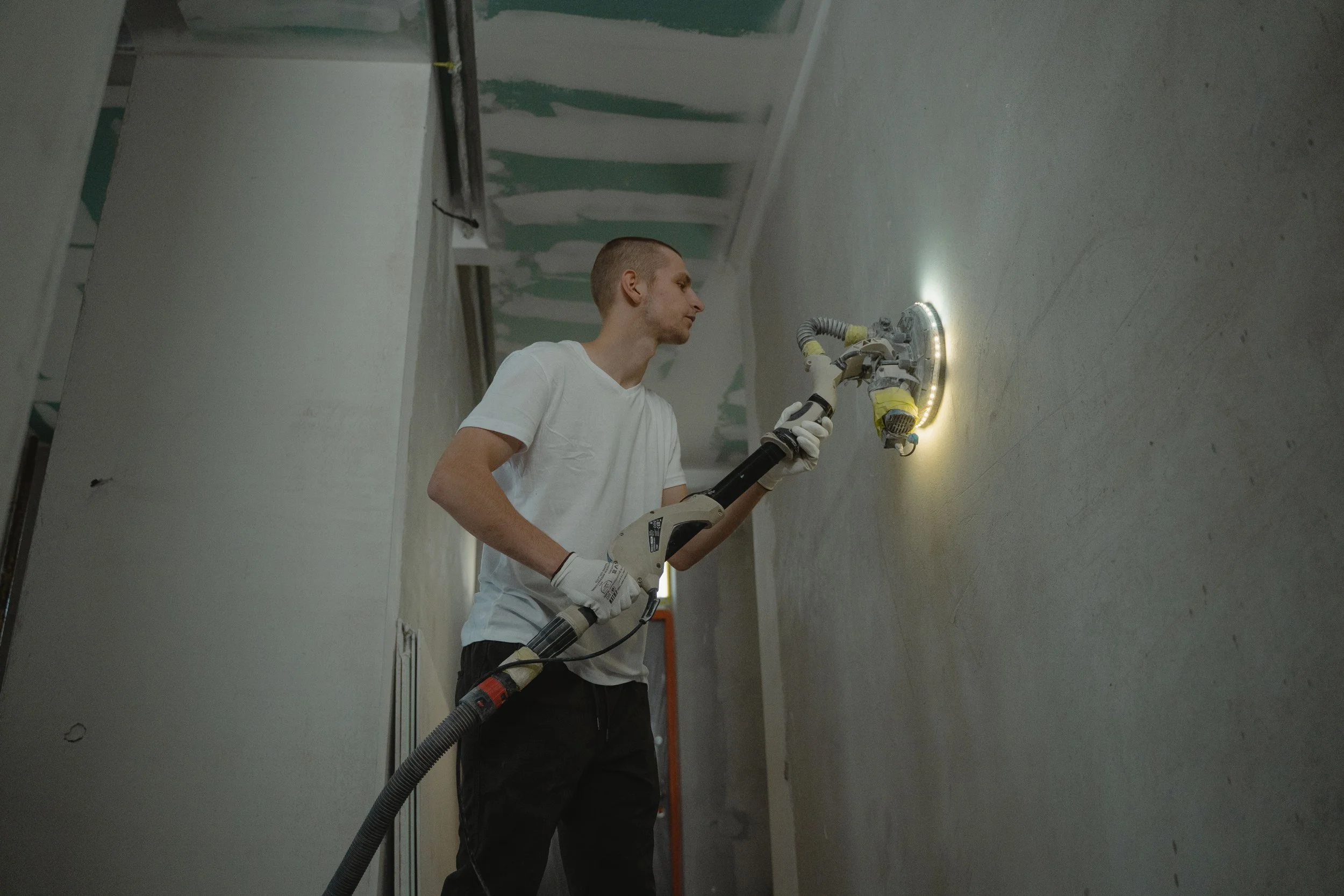 A man working on a wall, using a power sander, wearing a white t-shirt and gloves, in a room under construction with unfinished drywall ceiling.