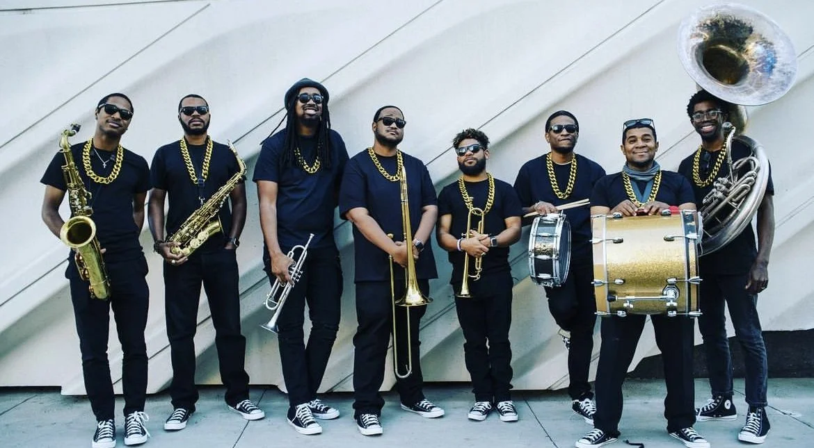 Eight musicians in coordinated black attire posing outdoors with instruments.