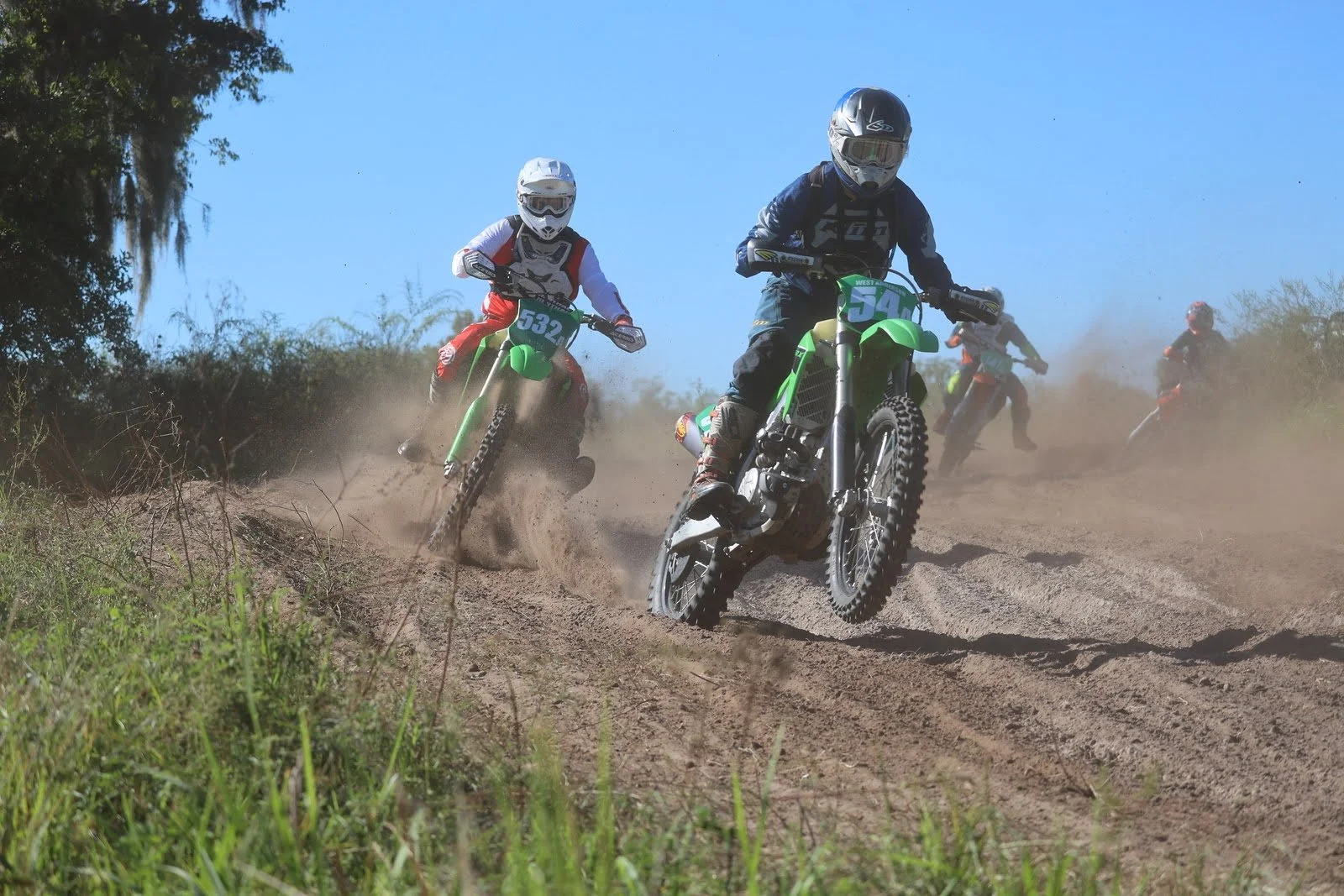 Group of motocross riders racing on a dirt track, kicking up dust under a clear blue sky.