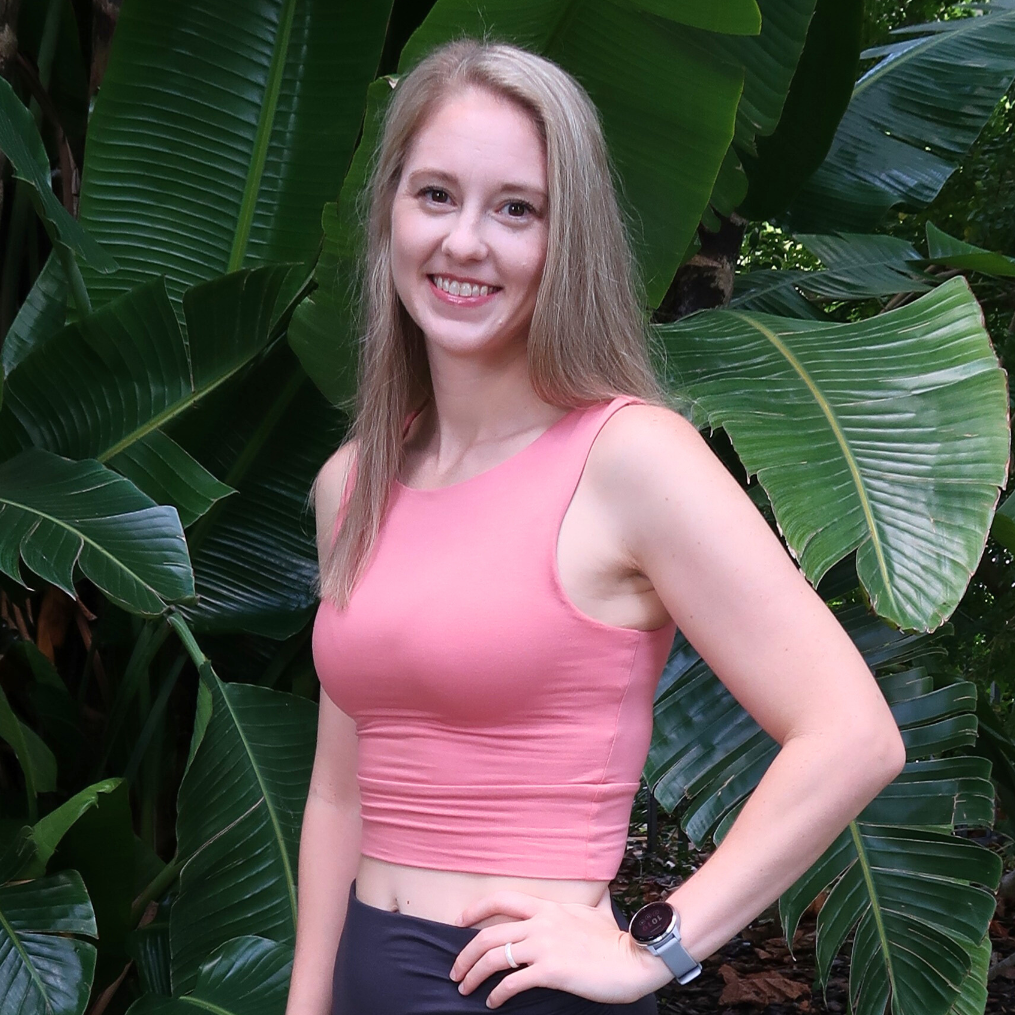 Close-up of a smiling woman with long blonde hair wearing a pink sleeveless top and a smartwatch, standing outdoors in front of large green tropical leaves.