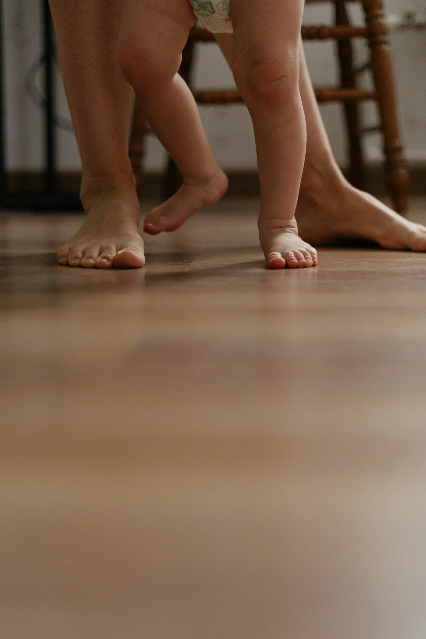 A close-up photo of an adult and a small child together on the floor, focusing on their legs and feet. The child appears to be standing or crawling next to the adult.