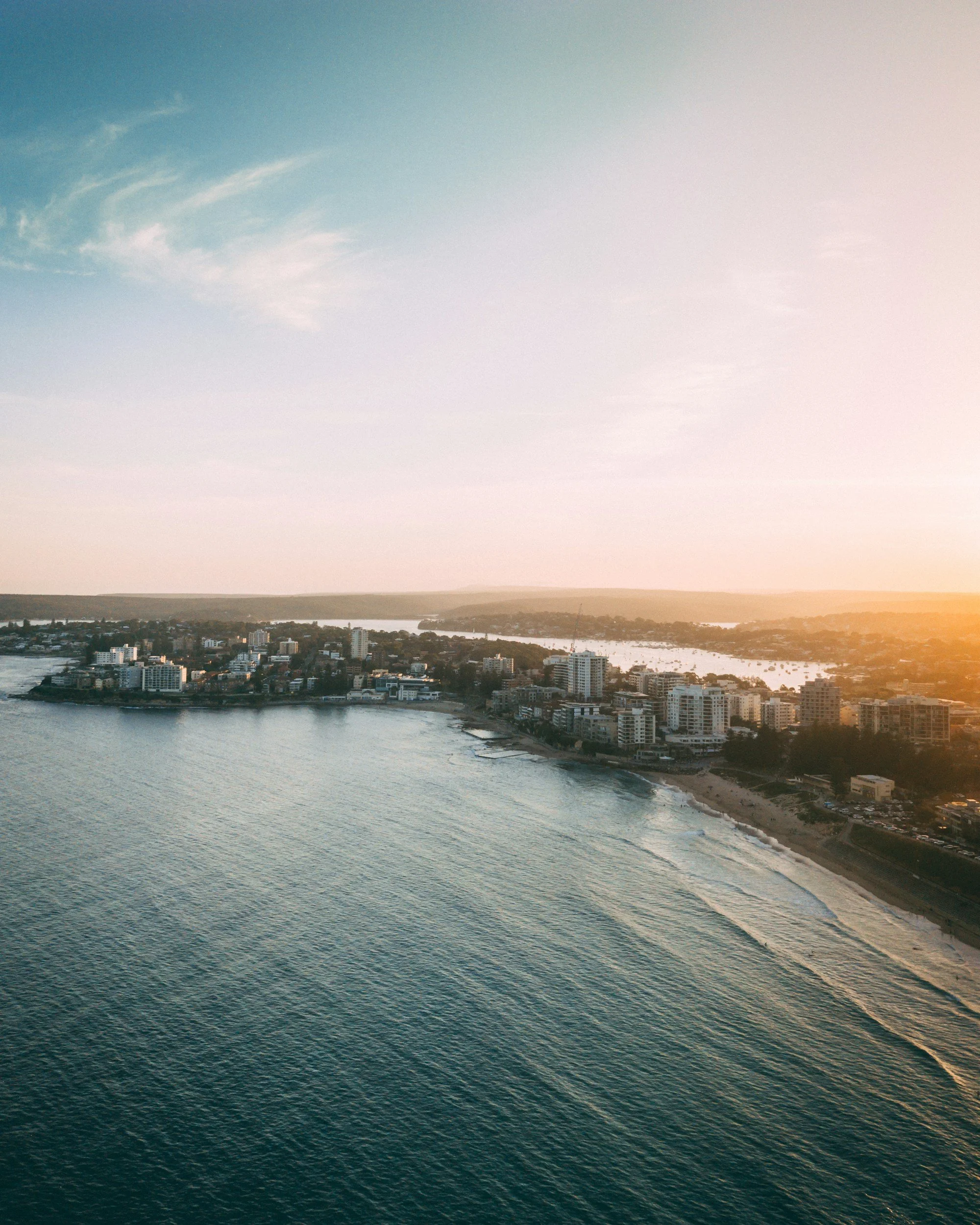 Aerial view of a coastal city with high-rise buildings along the shoreline at sunset, with a body of water in the foreground.