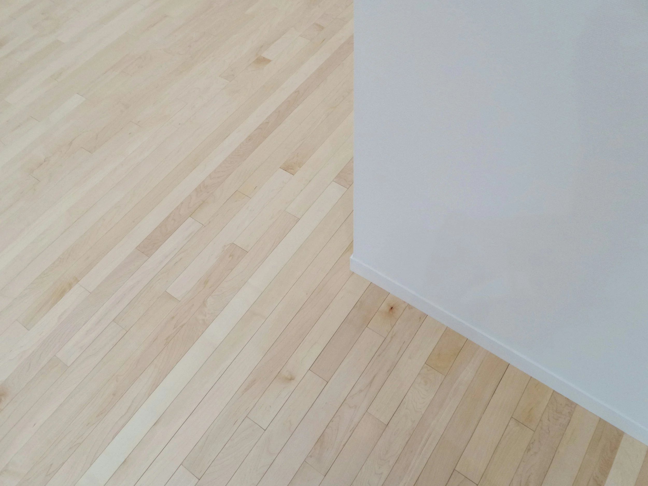 A corner of a room showing light wood flooring and a white wall with a baseboard.