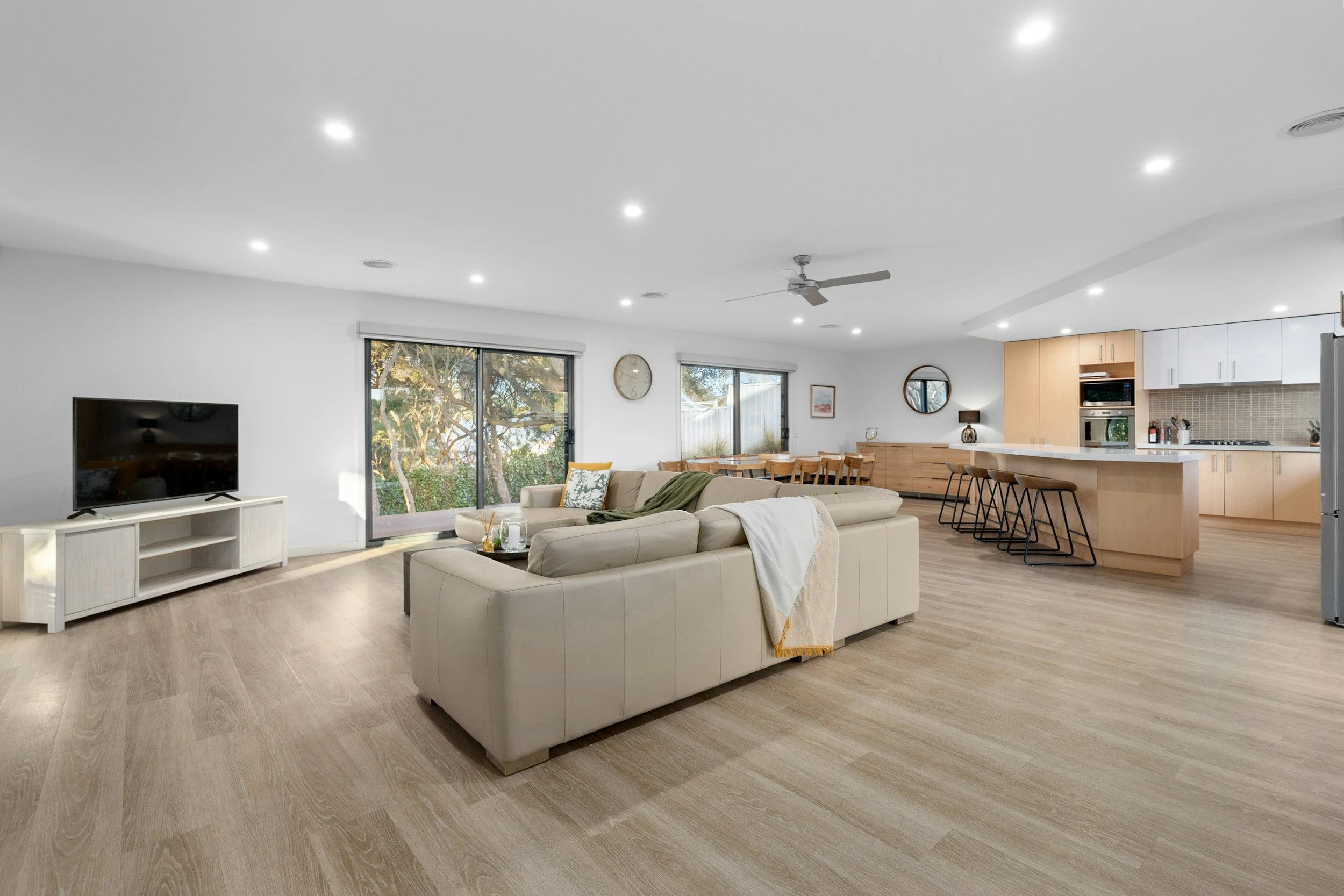Spacious open-plan living room and kitchen with wood flooring, white walls, large windows, and modern furniture.