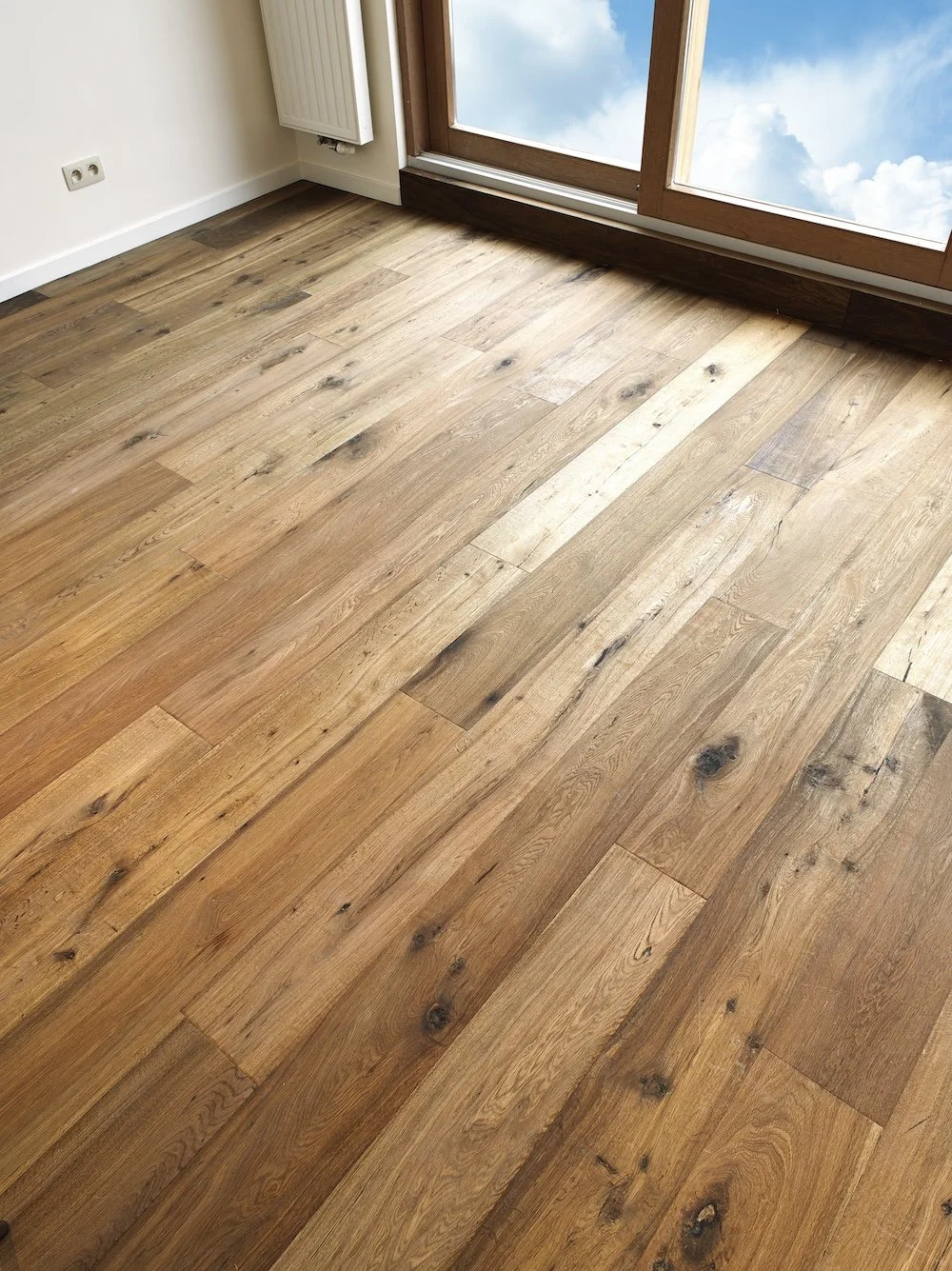 Wooden flooring in a room with a window showing a blue sky with clouds, a white radiator, and a power outlet on the wall.