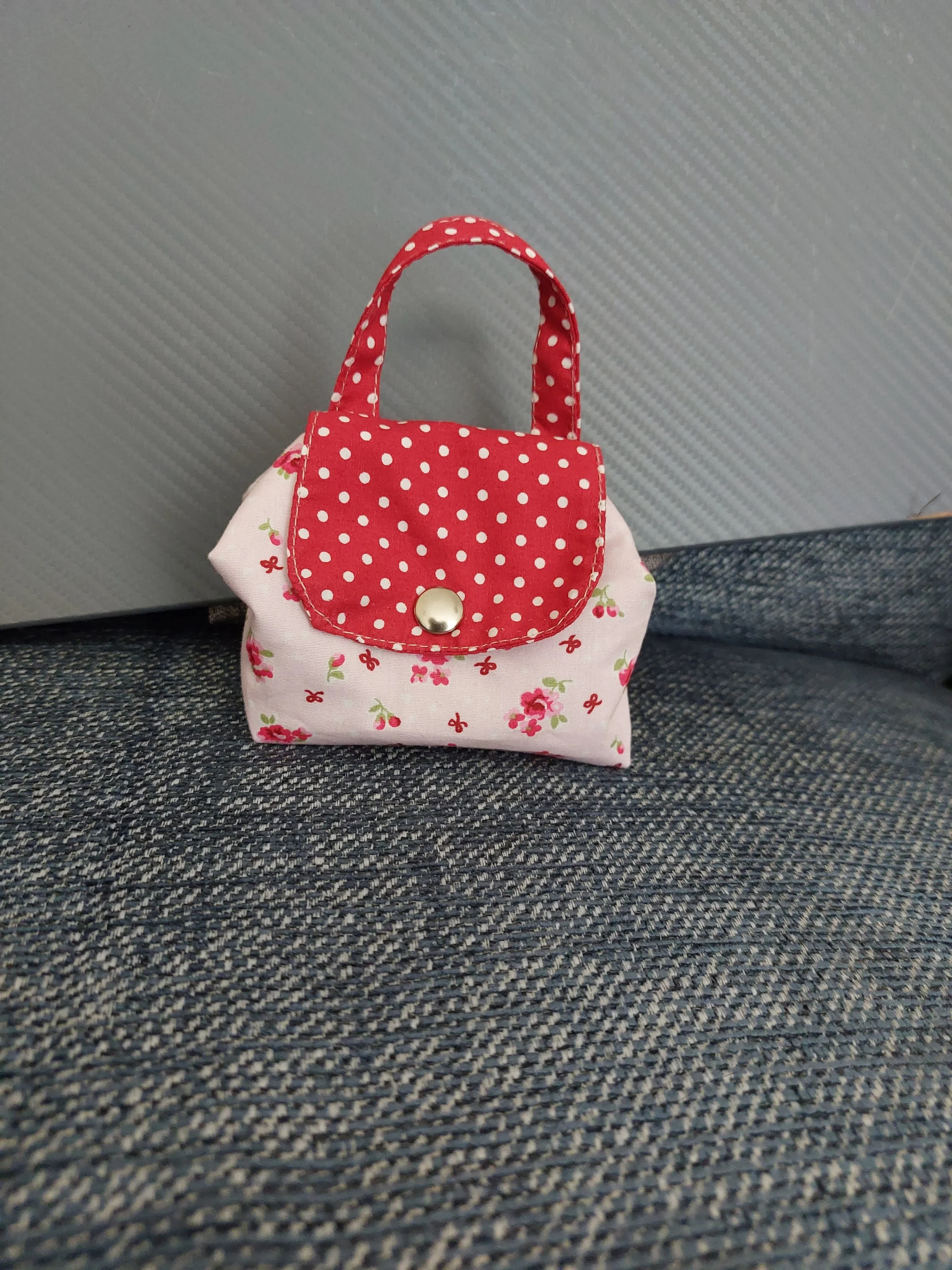 A small fabric bag with a red polka dot flap and handle, decorated with a white floral pattern, sitting on a gray and black textured surface.