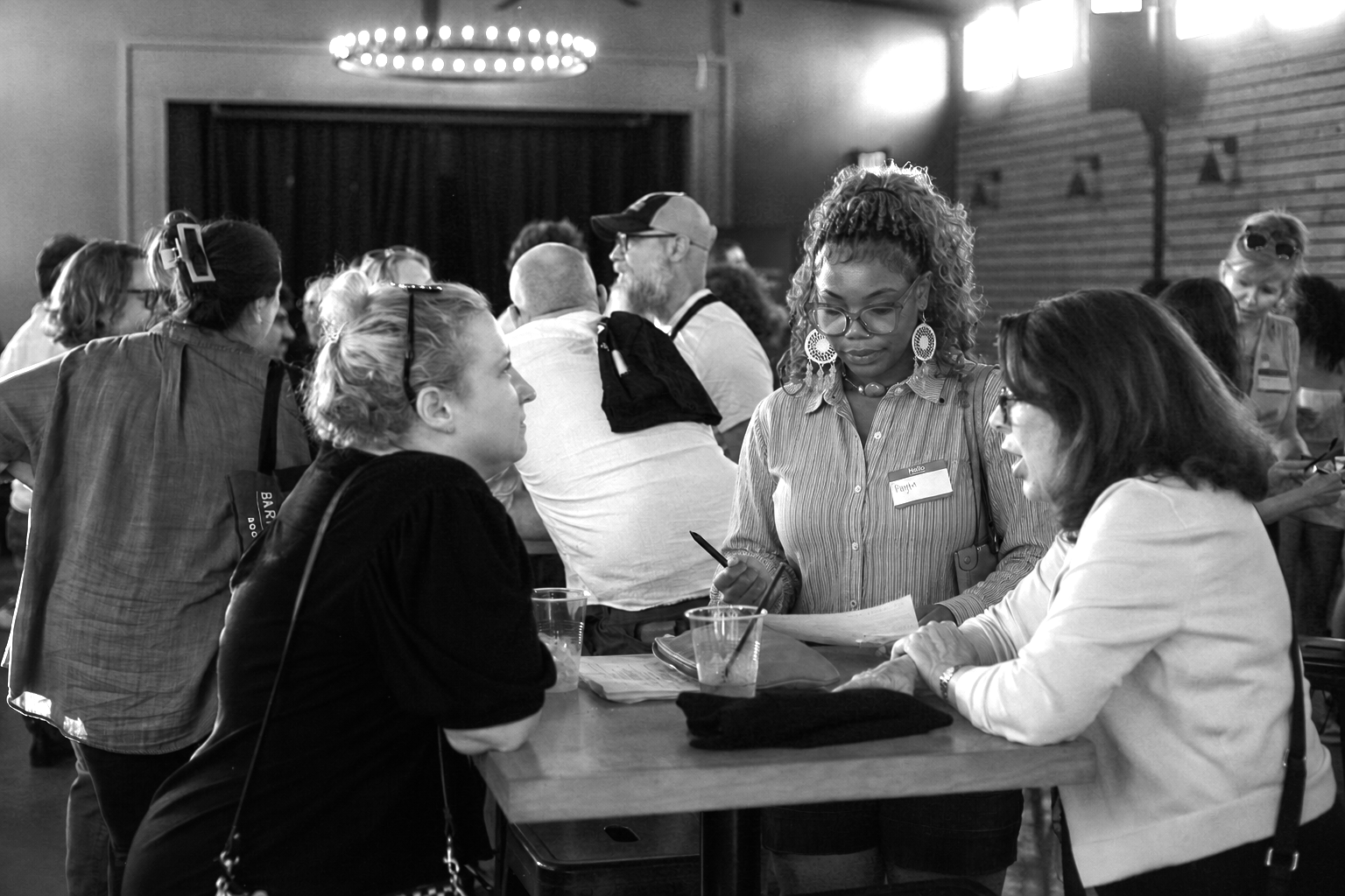 Three women engaged in conversation at a social event, with a gathering of people in the background.