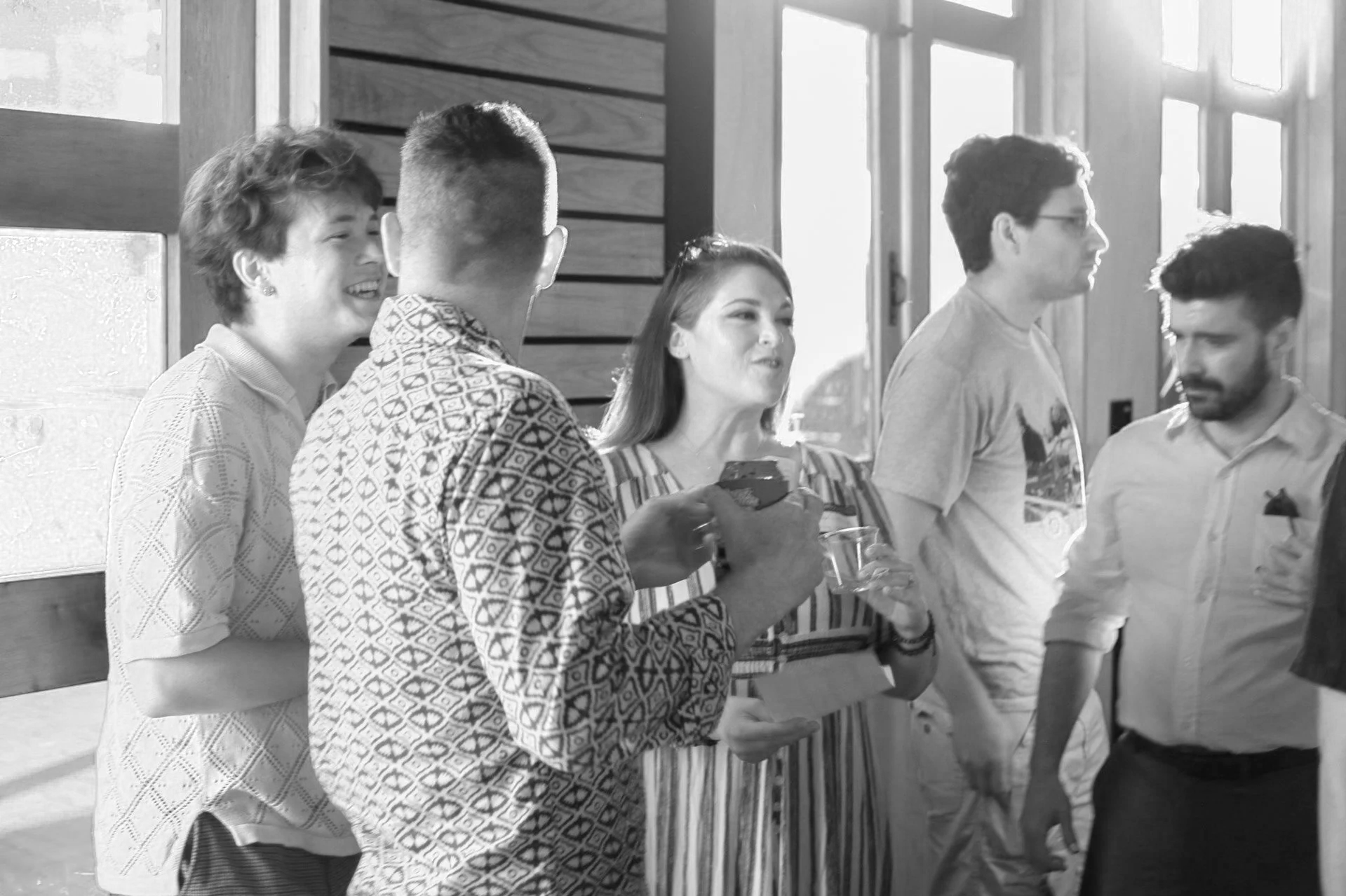 Group of people socializing indoors, some holding drinks, smiling, and engaging in conversation, with bright natural light coming through the windows.