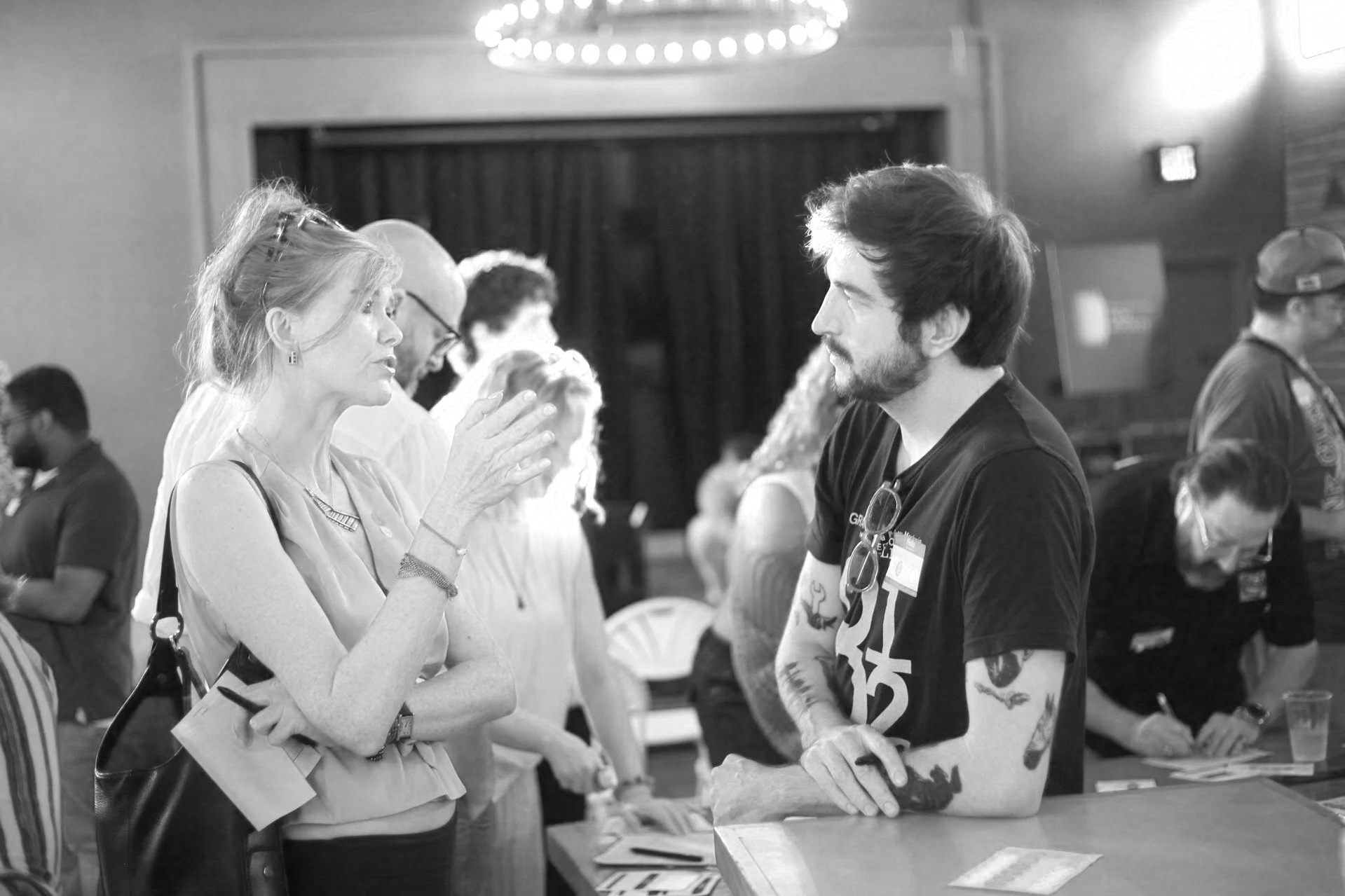 A woman and a man are engaged in conversation at a booth in an indoor event. The woman, with light hair and wearing a sleeveless top, gestures as she speaks. The man, with tattoos on his arms and wearing a T-shirt, listens attentively. There are other people in the background, some engaged in conversations or activities, and tables with papers.
