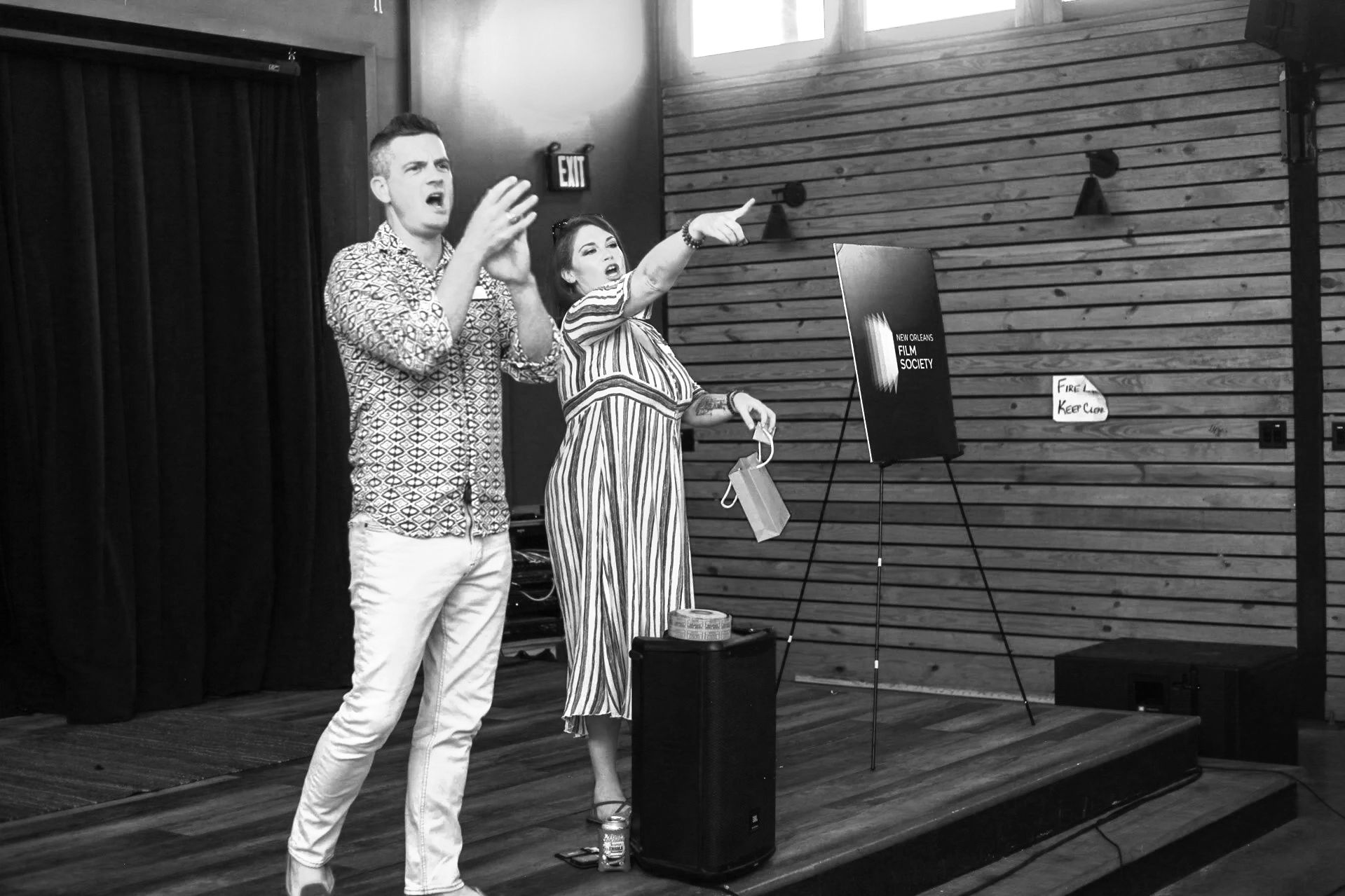 Two individuals, a man and a woman, are performing on a stage, gesturing with their hands. The man, on the left, has short hair and is wearing a patterned button-up shirt and white pants. The woman, on the right, has long hair and is wearing a striped dress, holding a small bag and pointing outward. They appear to be engaged in an animated discussion or presentation, with a sign for the 'New Orleans Film Society' on a stand, a small table with a can and other items, and a wooden-paneled wall in the background.